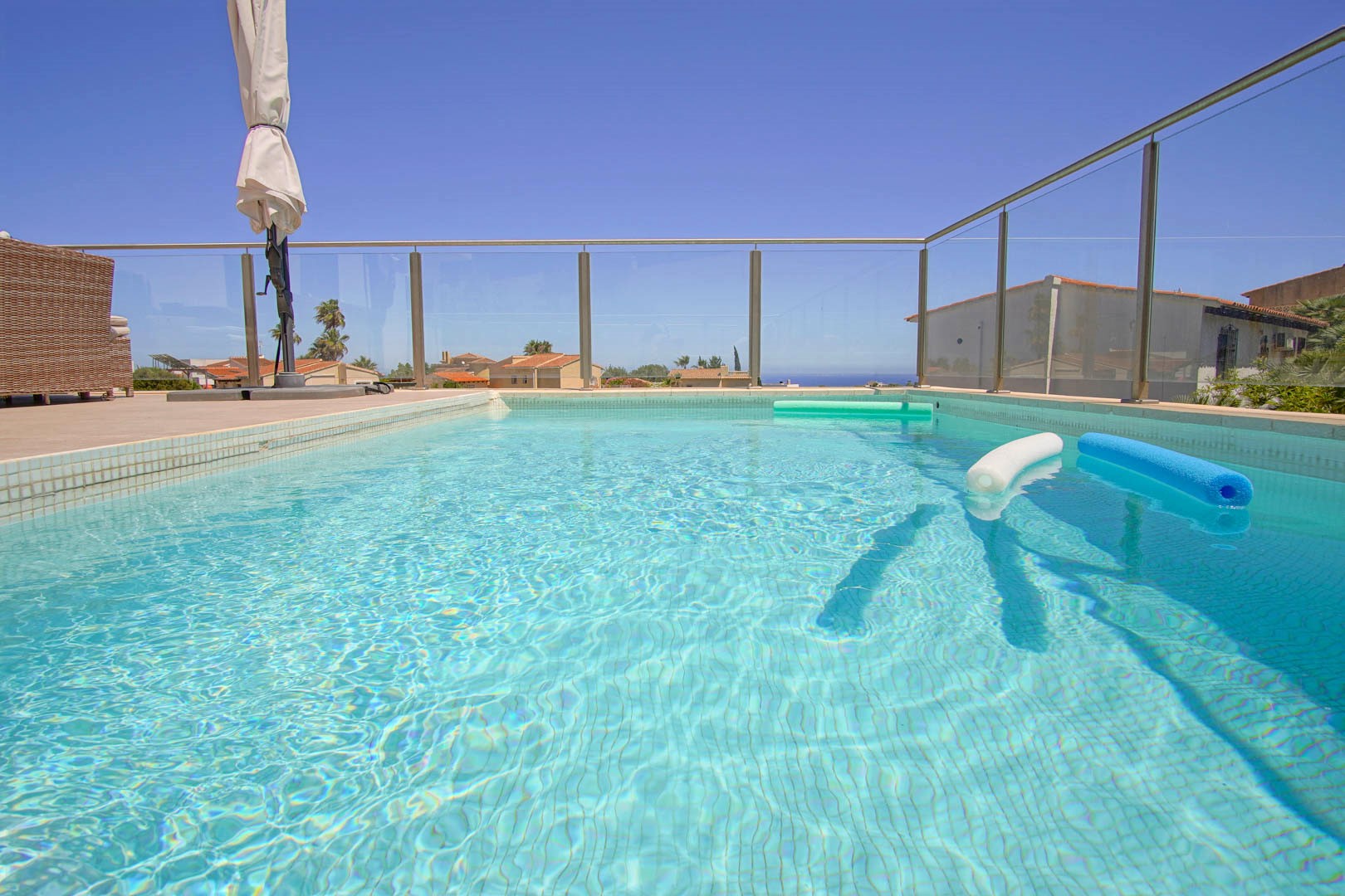 Piscina rectangular con revestimiento de gresite claro y barandilla de cristal de seguridad. La terraza ofrece vistas despejadas al mar y mobiliario de exterior en tonos neutros.
