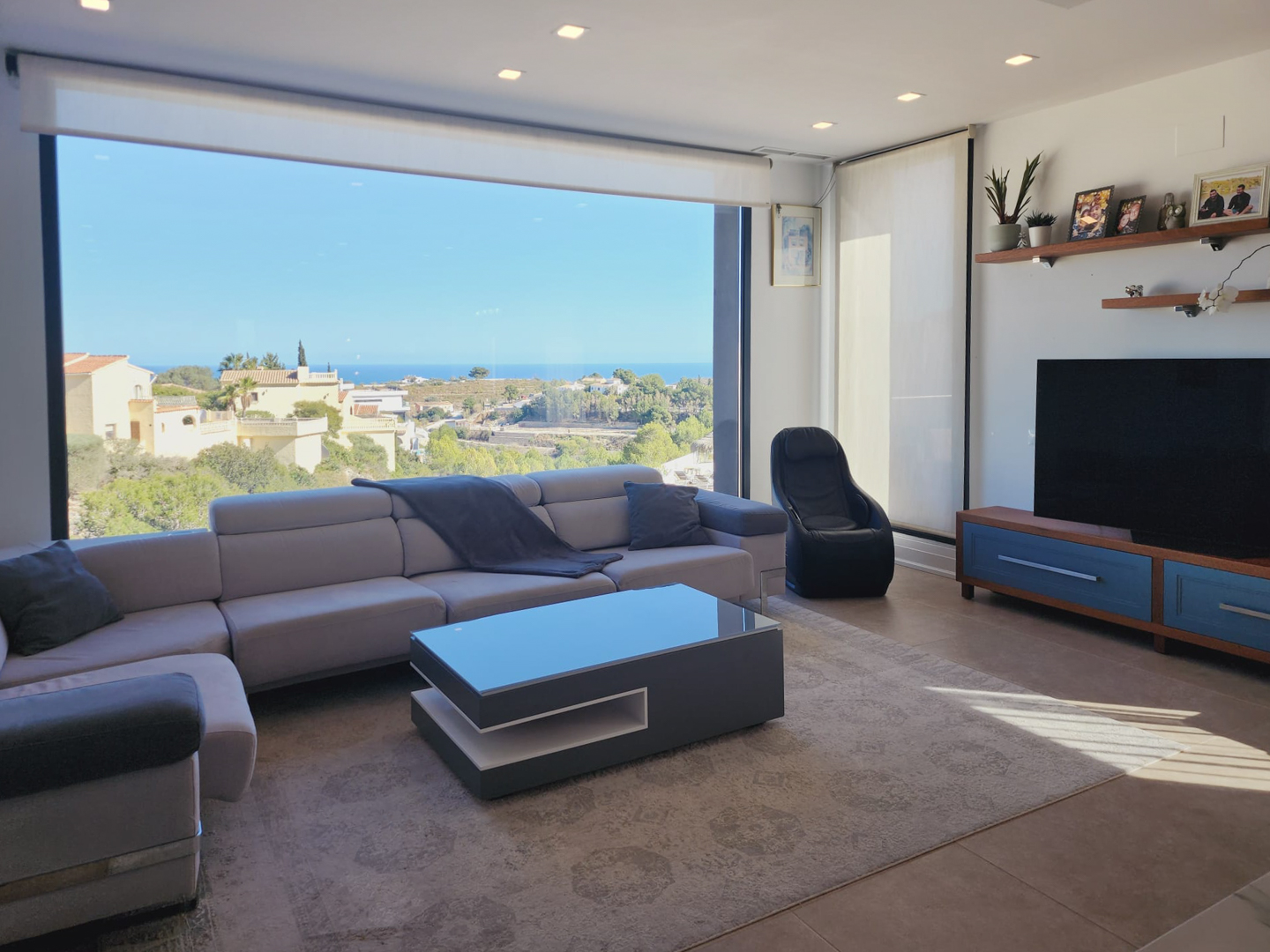 Salón moderno con ventanal panorámico al mar, sofá rinconera gris, mesa de centro de diseño y suelos cerámicos con iluminación empotrada.
