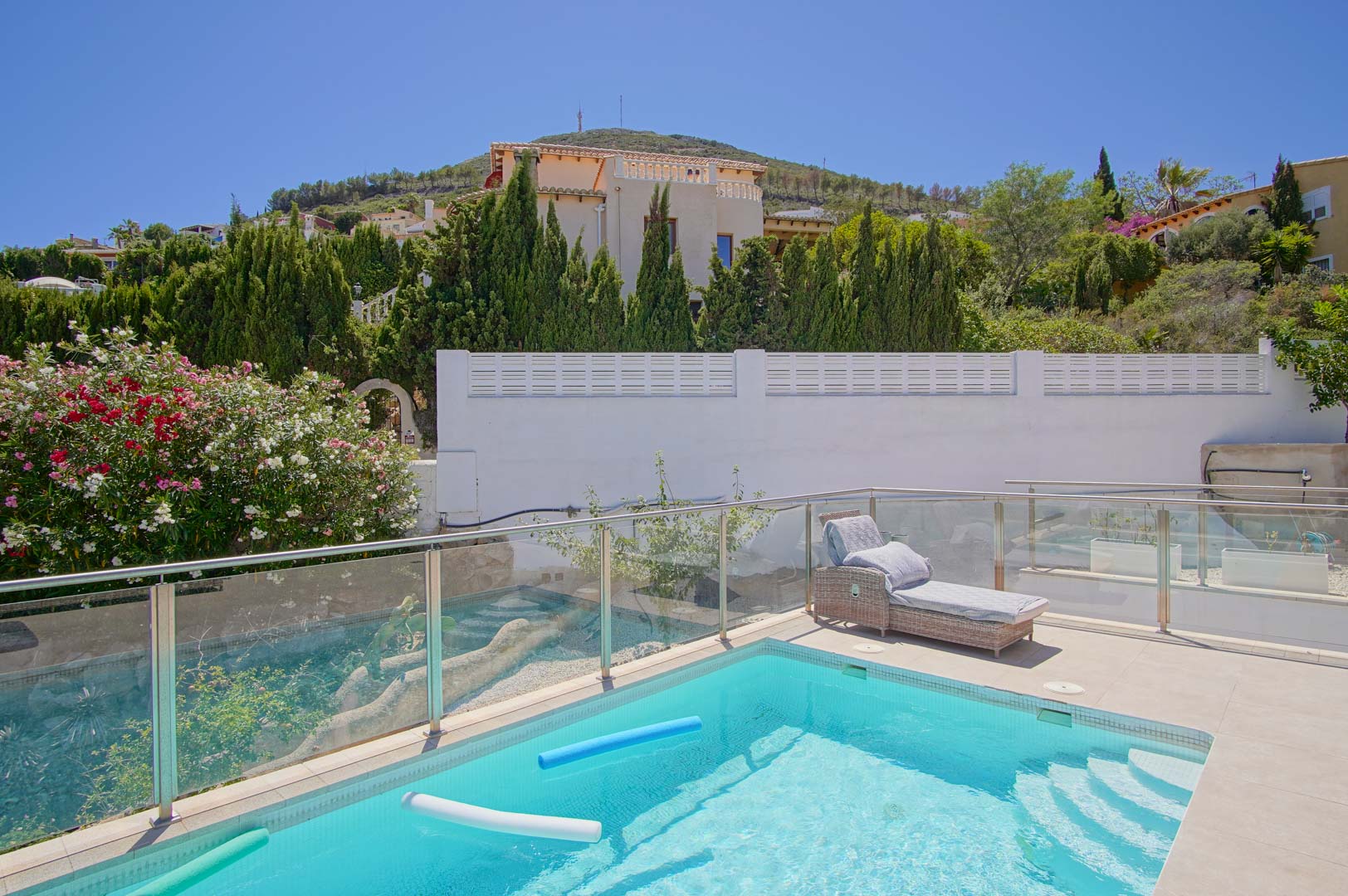 Terraza con piscina de aguas cristalinas, barandillas de vidrio y acero, y zona de descanso con tumbona de mimbre rodeada de adelfas y cipreses.