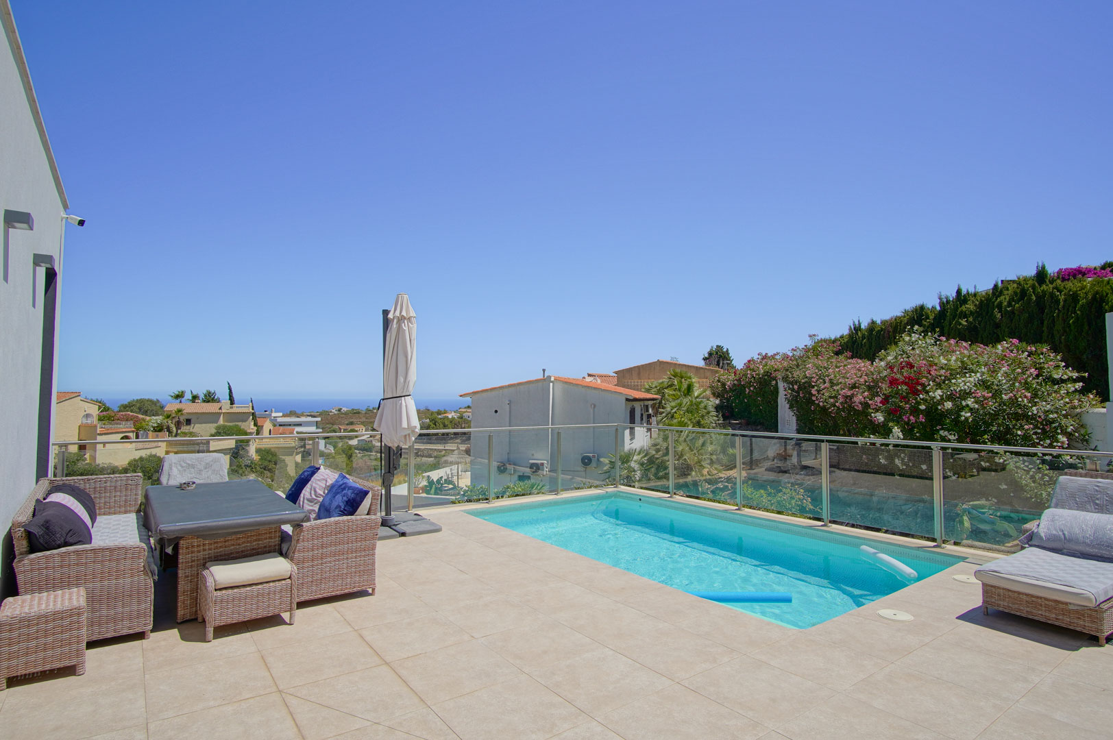 Terraza exterior con piscina rectangular, barandilla de cristal y mobiliario de ratán. Ofrece vistas despejadas al mar y orientación sur bajo un cielo despejado.