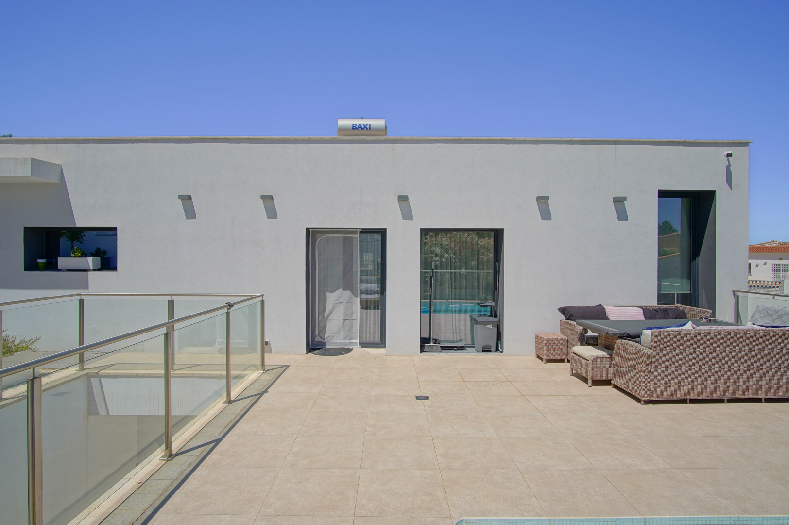 Terraza superior con pavimentos cerámicos en tonos arena, mobiliario de exterior de ratán y barandillas de cristal. Se observa el sistema de placas solares BAXI en la cubierta.