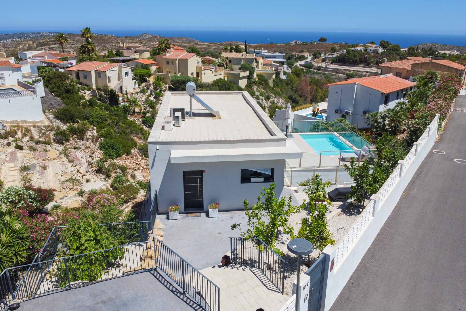 Vista aérea de la villa moderna con tejado plano, paneles solares y terraza con piscina vallada en cristal, destacando su ubicación con vistas al mar Mediterráneo.
