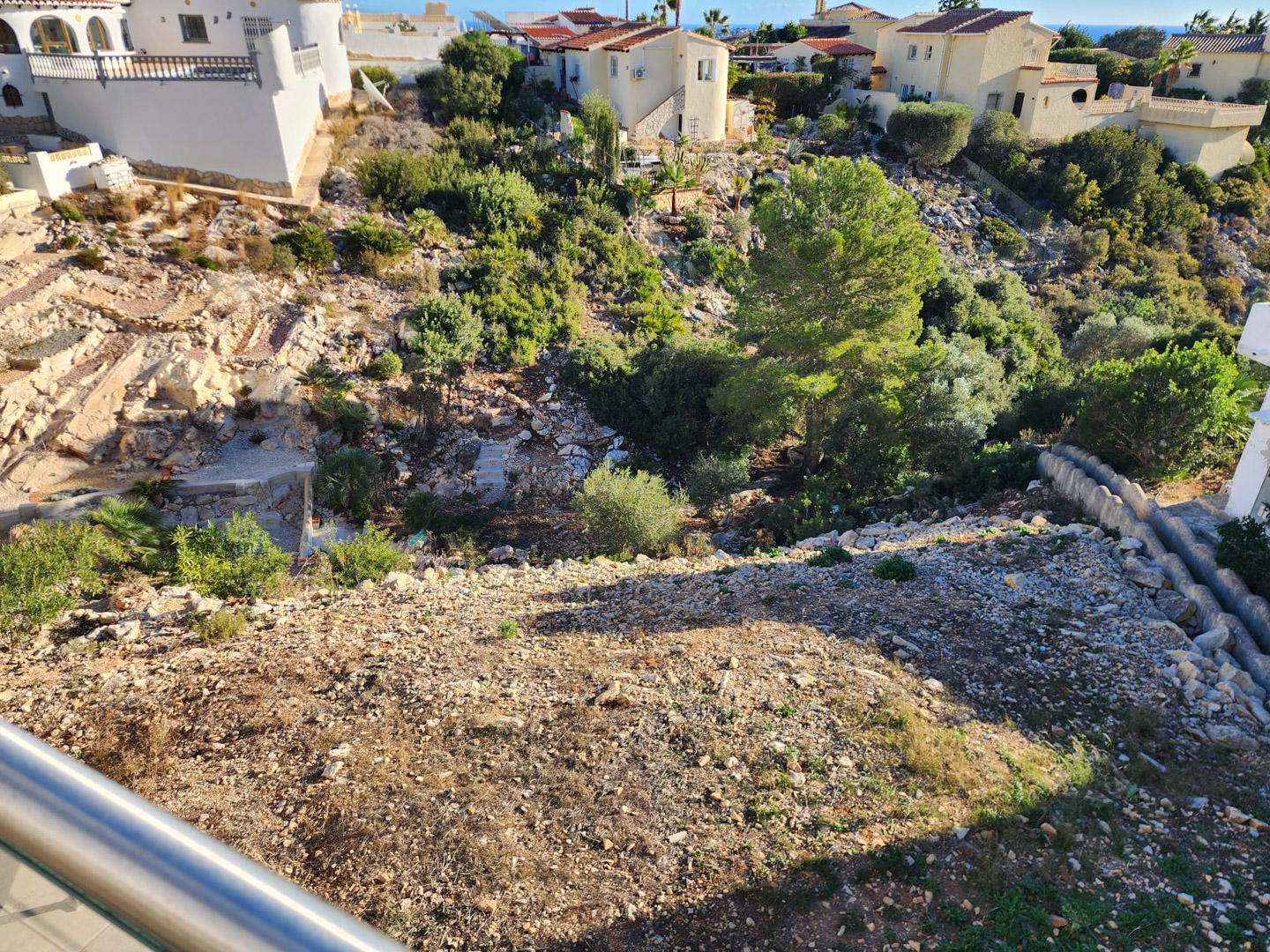 Vista desde el balcón hacia el terreno inclinado de la parcela, compuesto por suelo de grava, rocas naturales y vegetación autóctona mediterránea.