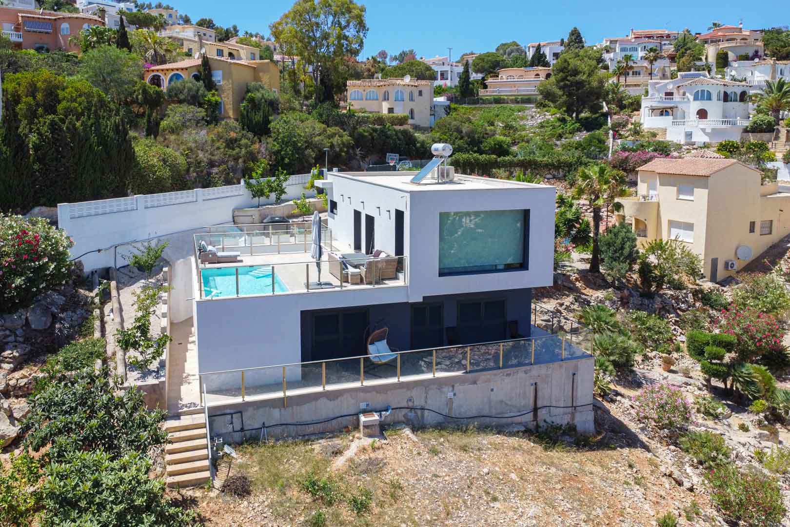 Vista exterior de la villa moderna con arquitectura cúbica, destacando la terraza superior con piscina, barandillas de cristal y paneles solares en la cubierta plana.