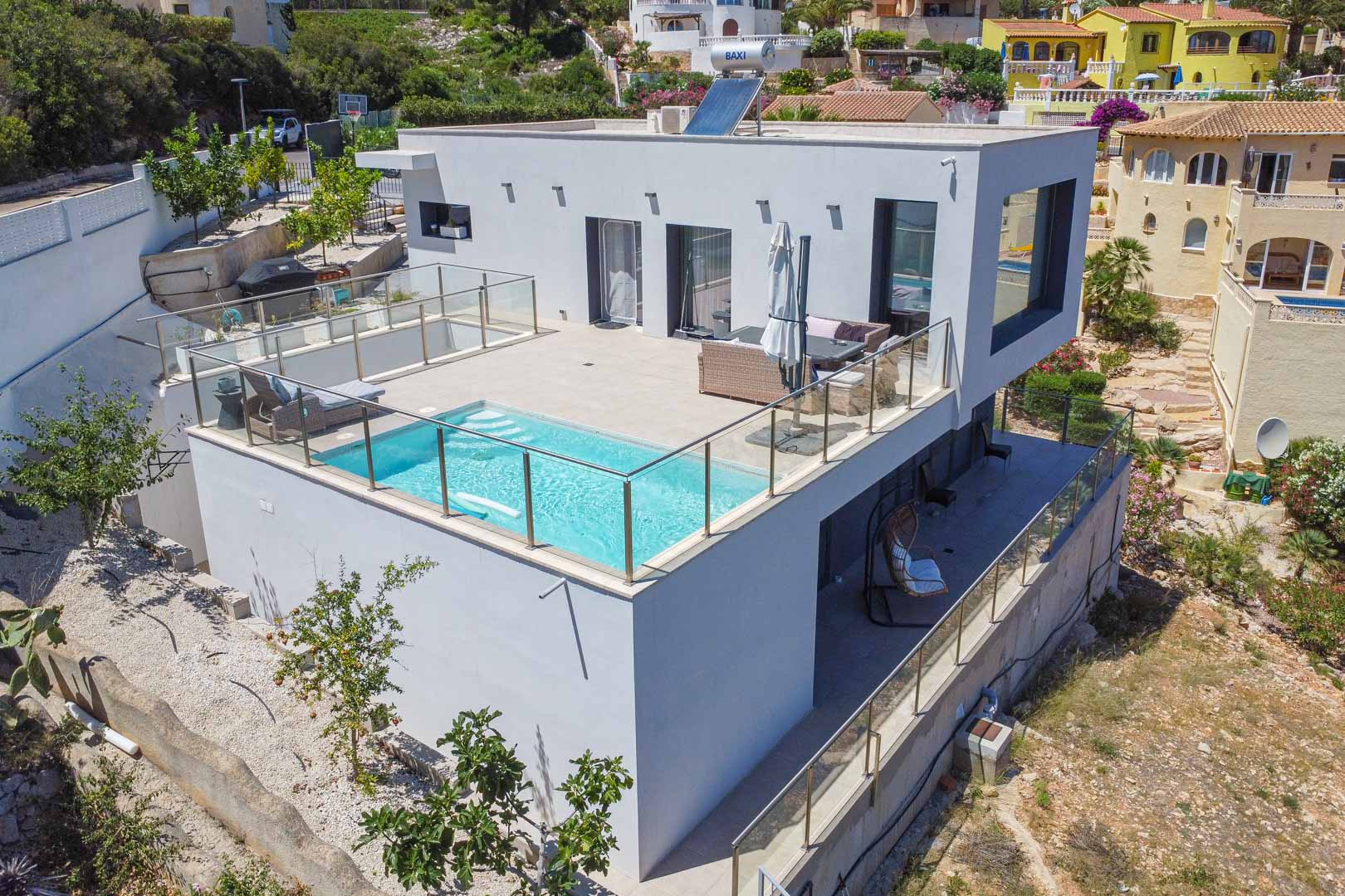 Vista exterior de la villa moderna con piscina integrada en la terraza superior, barandillas de cristal, paneles solares y una terraza inferior cubierta.