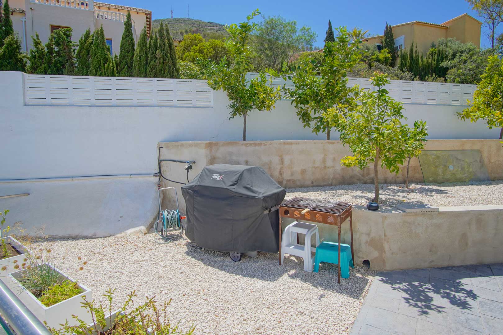 Zona exterior con grava blanca, árboles frutales y área de barbacoa. El espacio incluye un muro perimetral blanco y vistas a la vegetación circundante.