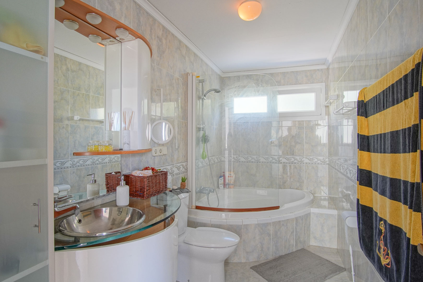 Baño completo con bañera de esquina, mampara de cristal y lavabo de acero inoxidable sobre encimera de vidrio. Paredes alicatadas con cenefa decorativa y luz natural.