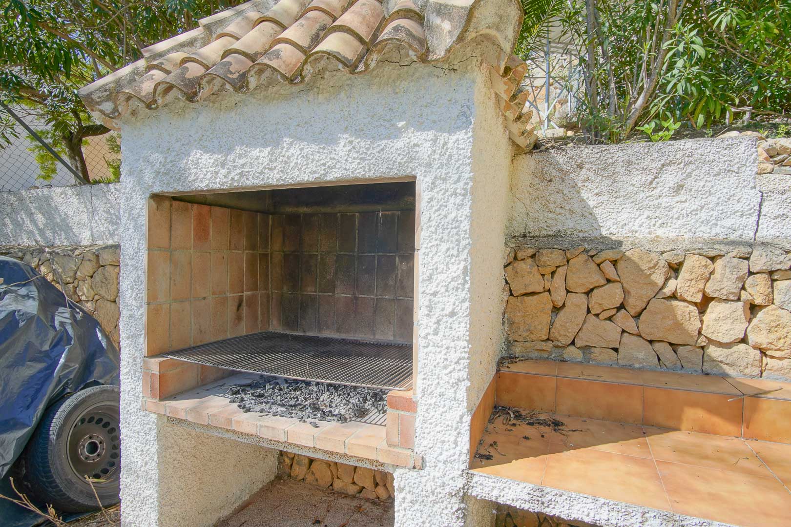 Barbacoa de obra con tejadillo de teja cerámica y paredes de estuco blanco, integrada junto a un muro de piedra natural y encimera lateral revestida en gres.