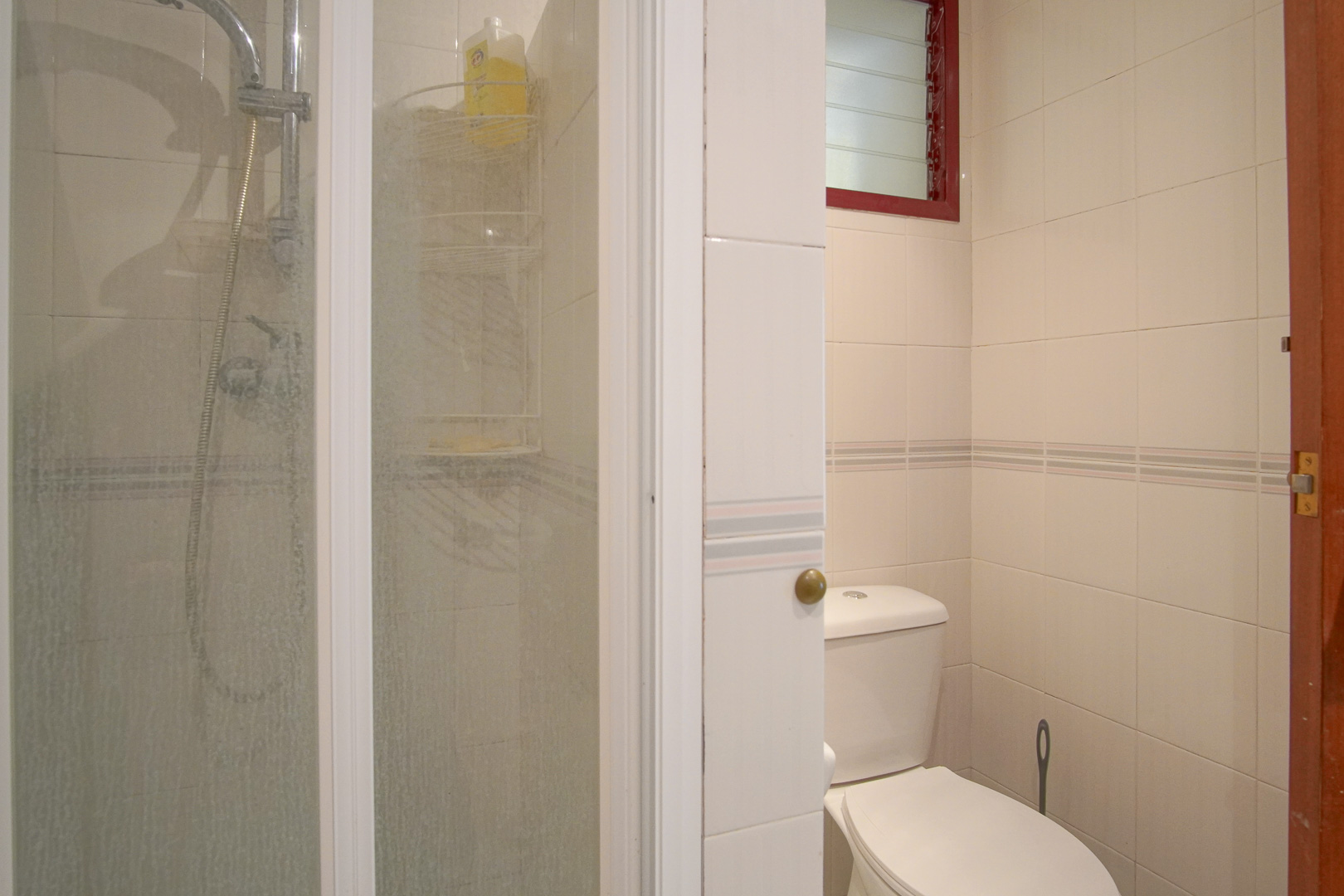 Cuarto de baño funcional con paredes alicatadas en blanco, ducha con mampara de cristal, inodoro estándar y ventana de ventilación superior con marco rojo.