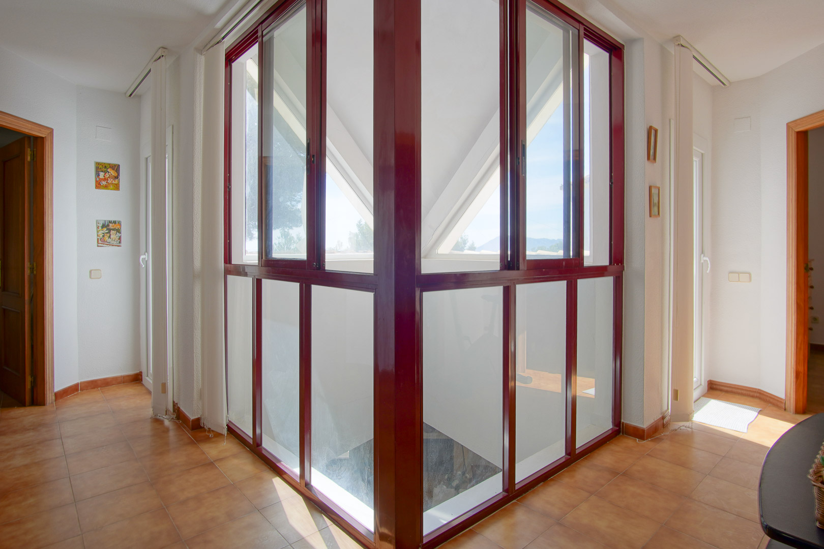 Distribuidor en planta superior con cerramiento acristalado de carpintería granate, suelos de gres rústico y abundante luz natural que conecta las estancias.
