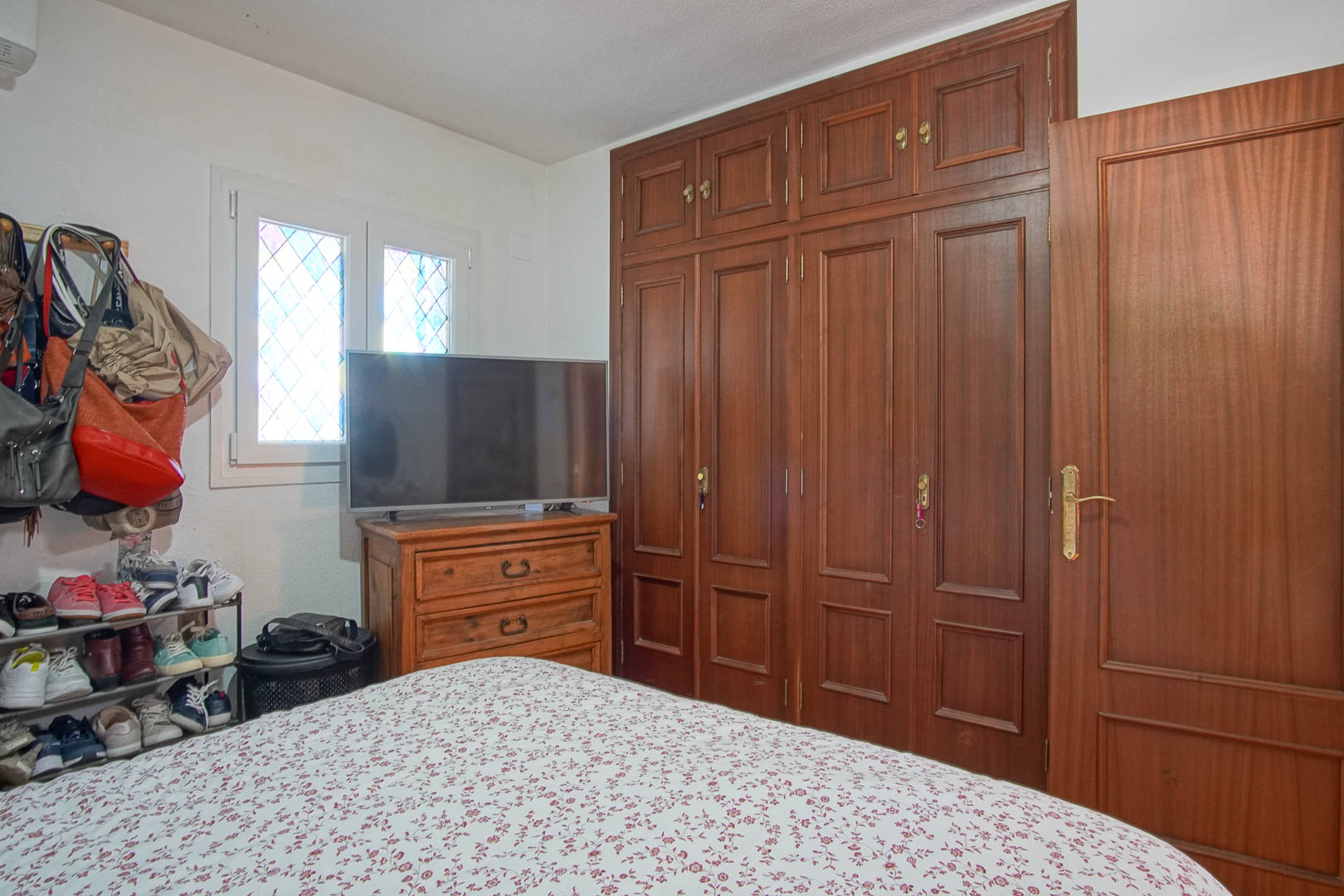 Dormitorio con amplio armario empotrado de madera oscura, cómoda a juego con televisor y ventana de cristal emplomado que aporta luz natural.