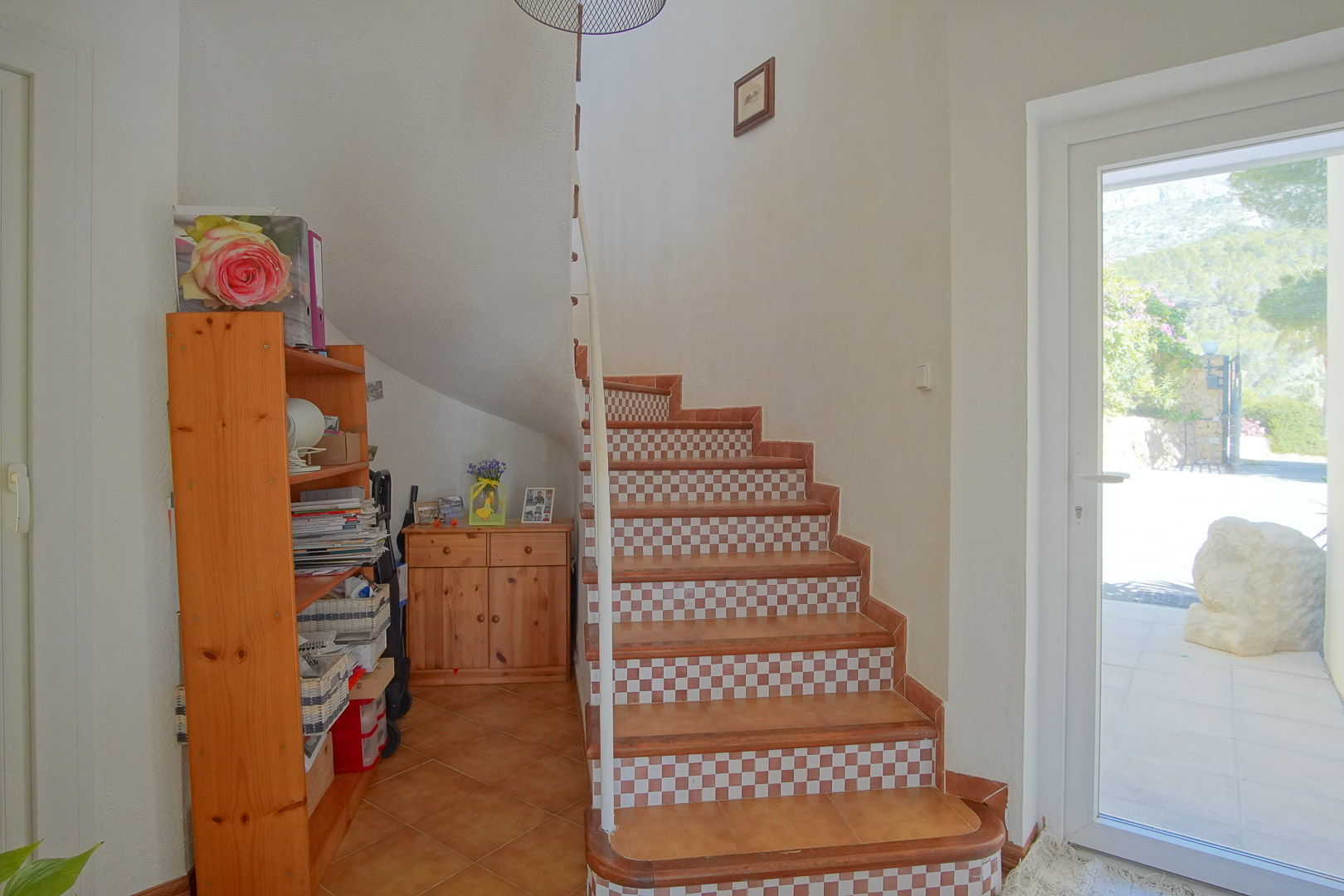 Escalera interior con peldaños de cerámica y contrahuellas en damero, mobiliario de pino y acceso directo al exterior mediante puerta acristalada.