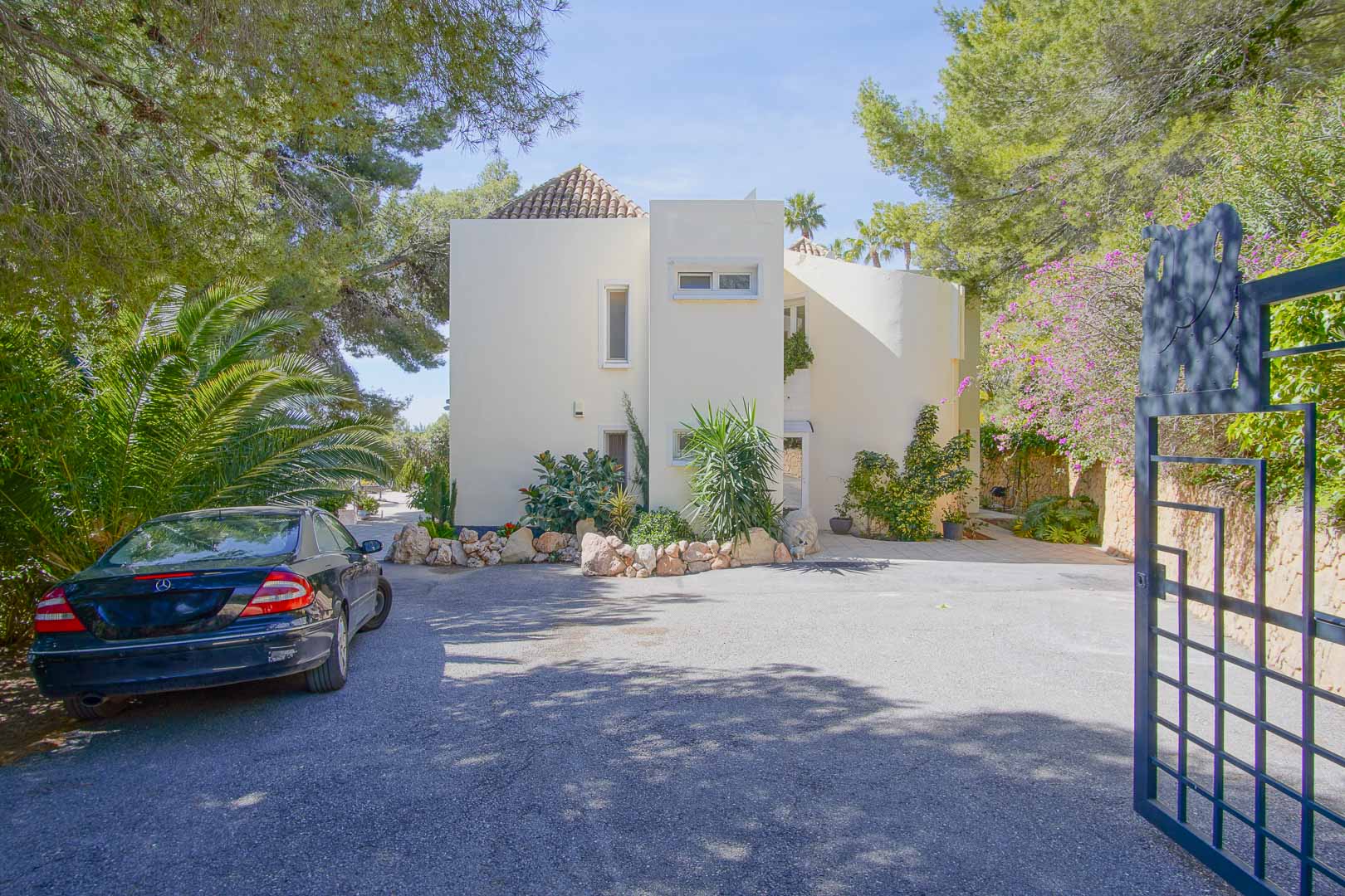 Fachada principal de la villa con arquitectura mediterránea, zona de aparcamiento privada y entrada flanqueada por pinos y vegetación autóctona en la Urbanización Santa Clara.