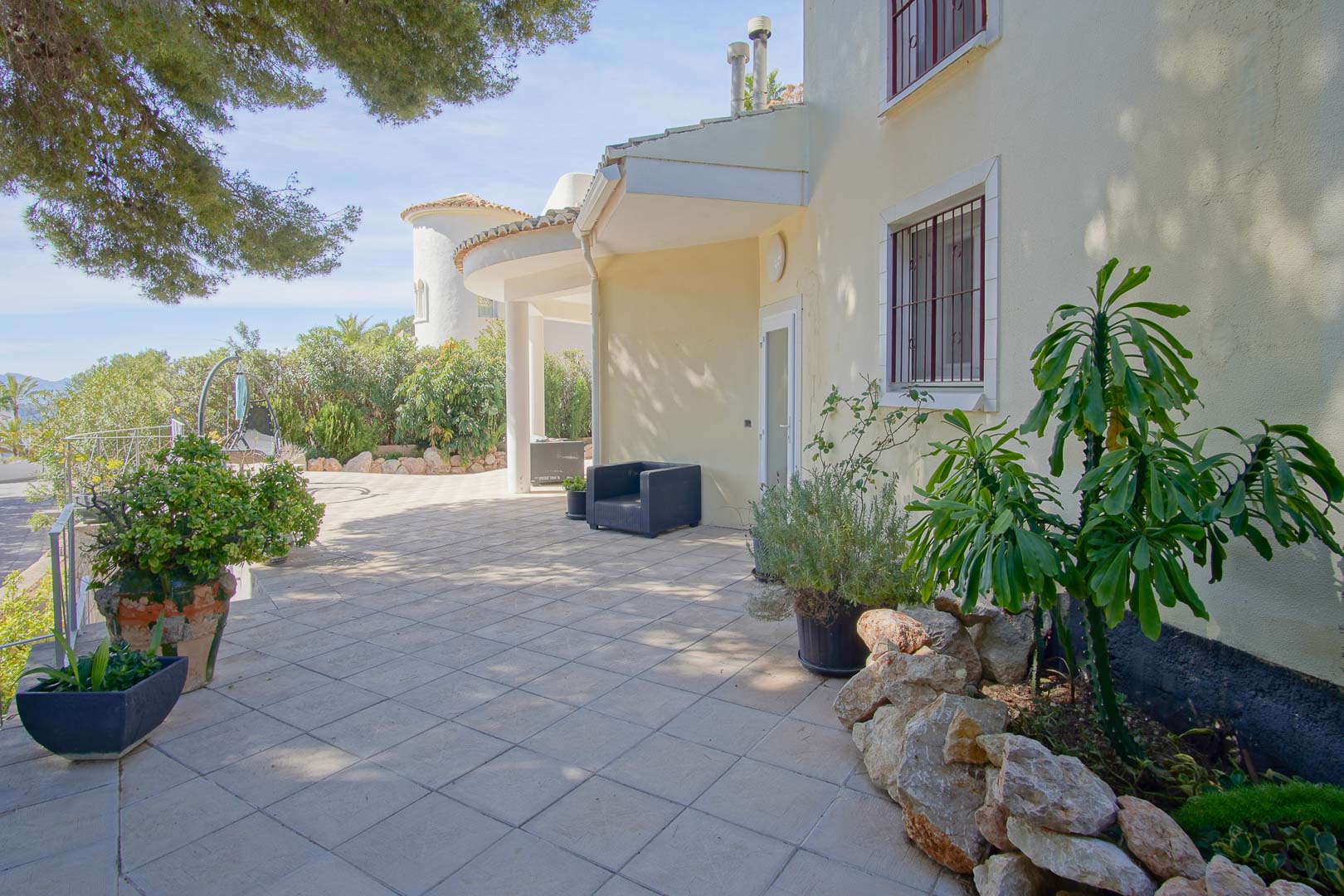 Patio exterior pavimentado con baldosas claras, rodeado de plantas mediterráneas y una fachada color crema con ventanas protegidas por rejas.