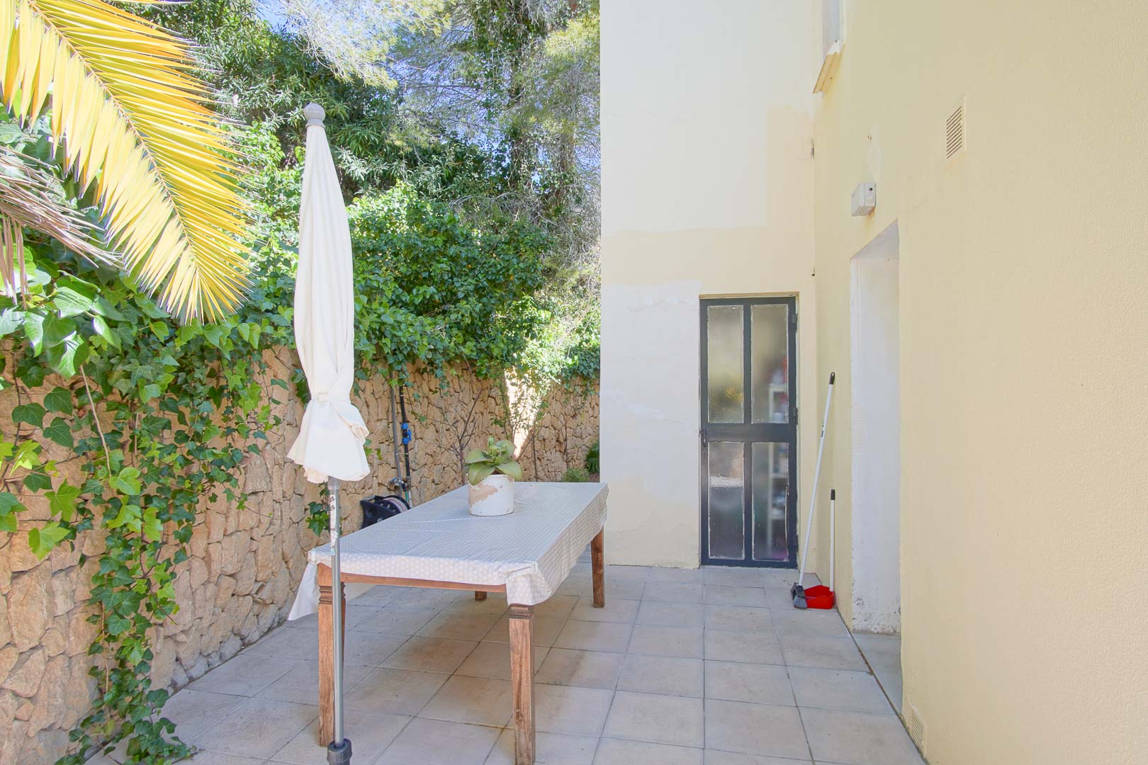 Patio exterior pavimentado junto a un muro de piedra natural con hiedra, equipado con mesa de comedor y sombrilla en un entorno privado y arbolado.