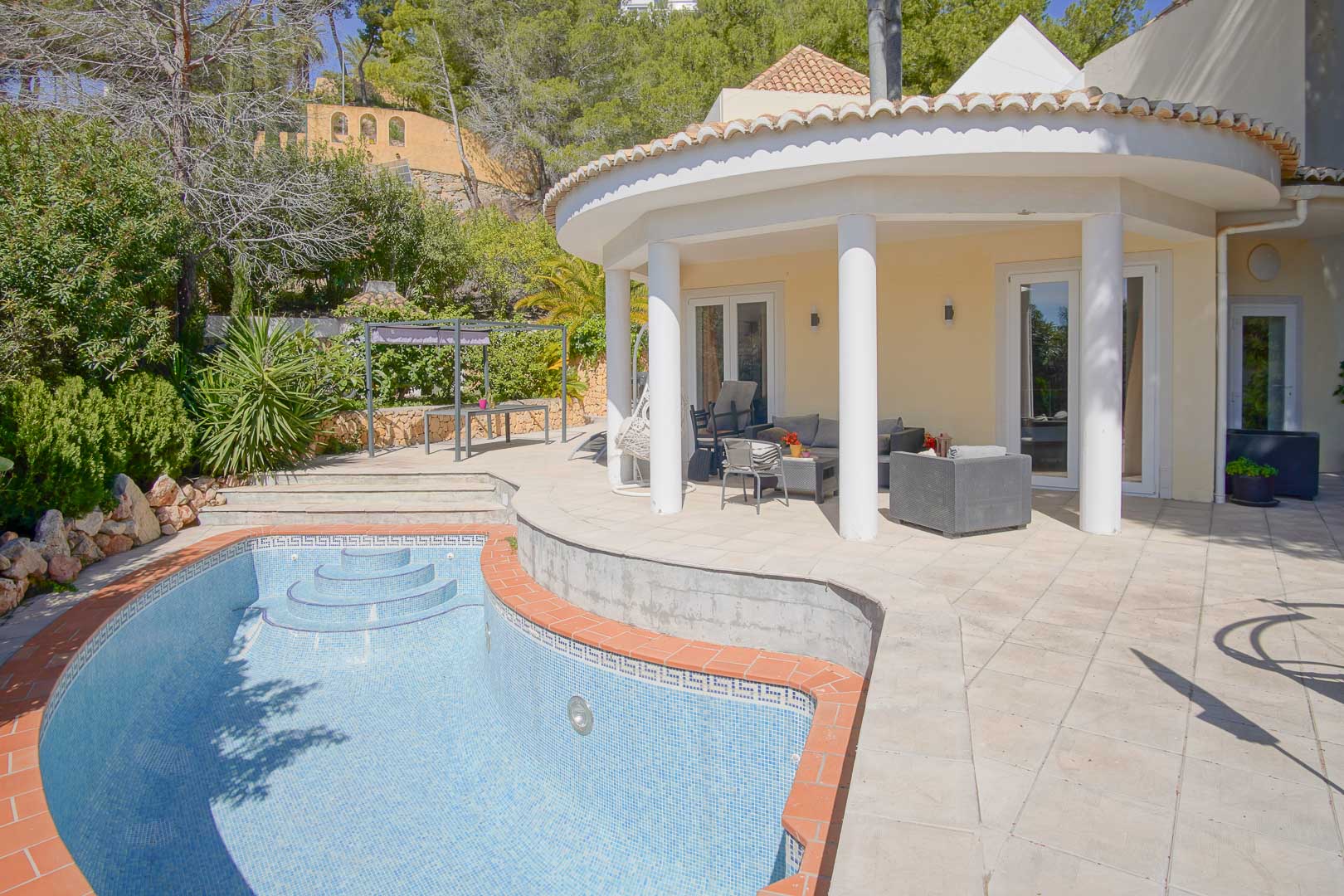 Porche semicircular con columnas blancas frente a una piscina privada con borde de terracota, rodeado de una terraza de piedra y vegetación mediterránea.