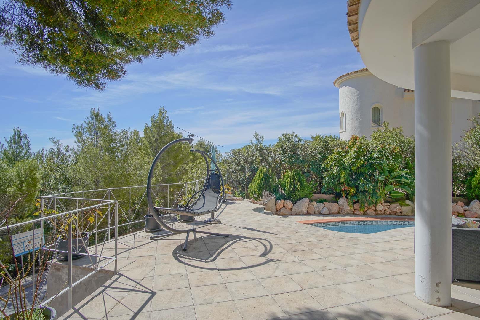 Terraza exterior pavimentada con sillón colgante, acceso a la piscina con cenefa griega y vistas a la vegetación mediterránea bajo un cielo despejado.
