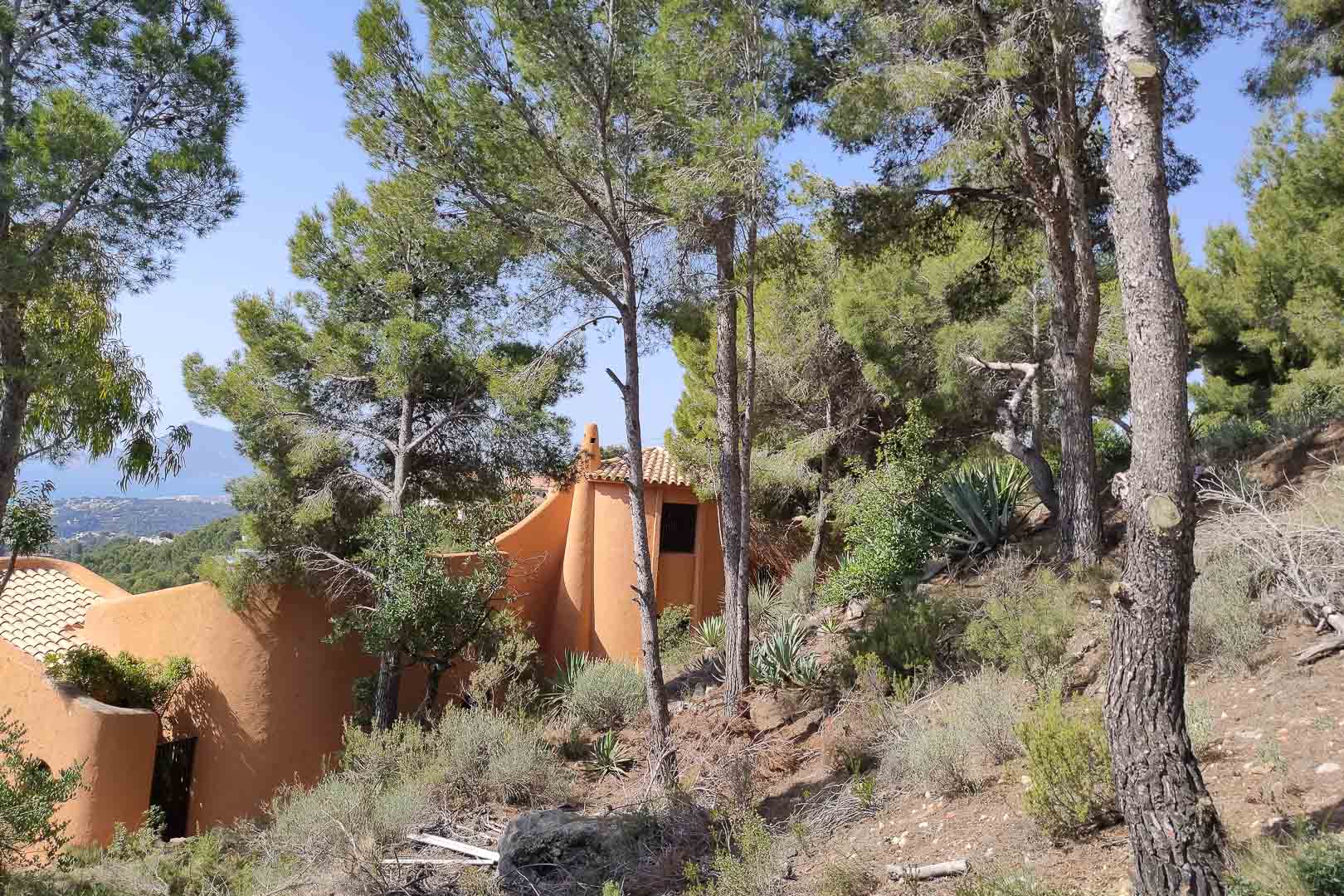 Vista exterior de la arquitectura orgánica en tonos terracota, rodeada de un denso bosque de pinos y vegetación mediterránea autóctona con vistas al mar.