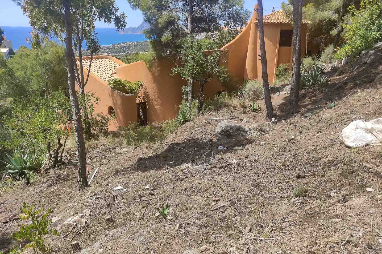 Vista exterior de la villa con arquitectura orgánica y fachadas en tonos tierra, integrada en una parcela elevada con pinos y vistas panorámicas al mar Mediterráneo.