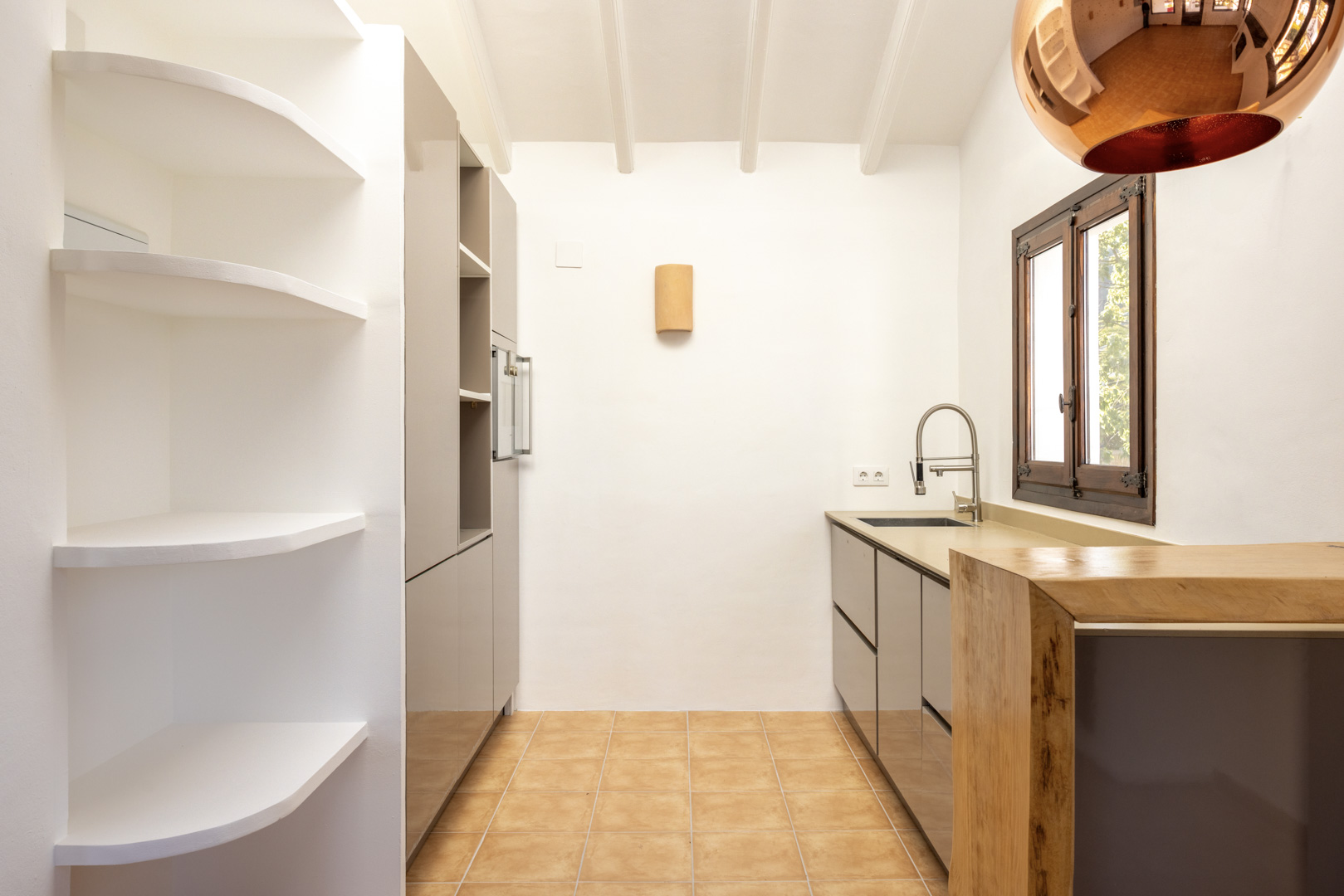 Cocina luminosa con estantes de obra blancos, armarios grises y encimera de madera. Suelo de baldosas rústicas.