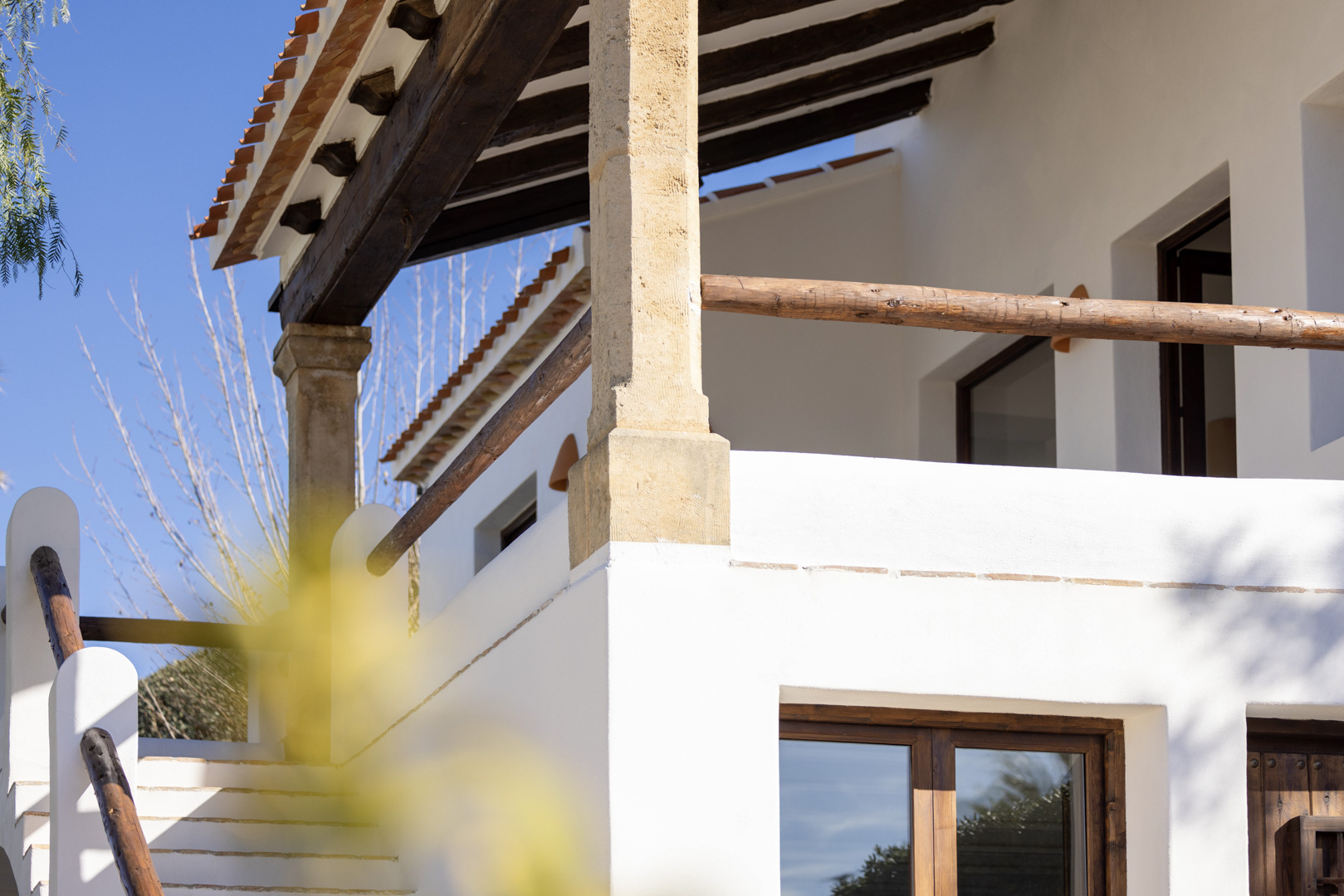 Detalle arquitectónico de la villa: porche con vigas de madera y barandilla rústica, escalera exterior de piedra blanca.
