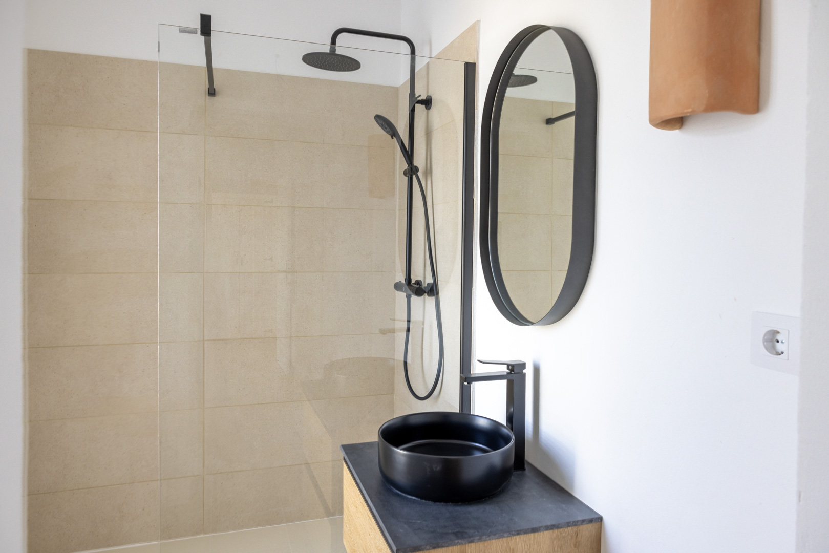 Detalle de baño con ducha de pared de azulejos beige, lavabo negro sobre encimera de pizarra y espejo ovalado.
