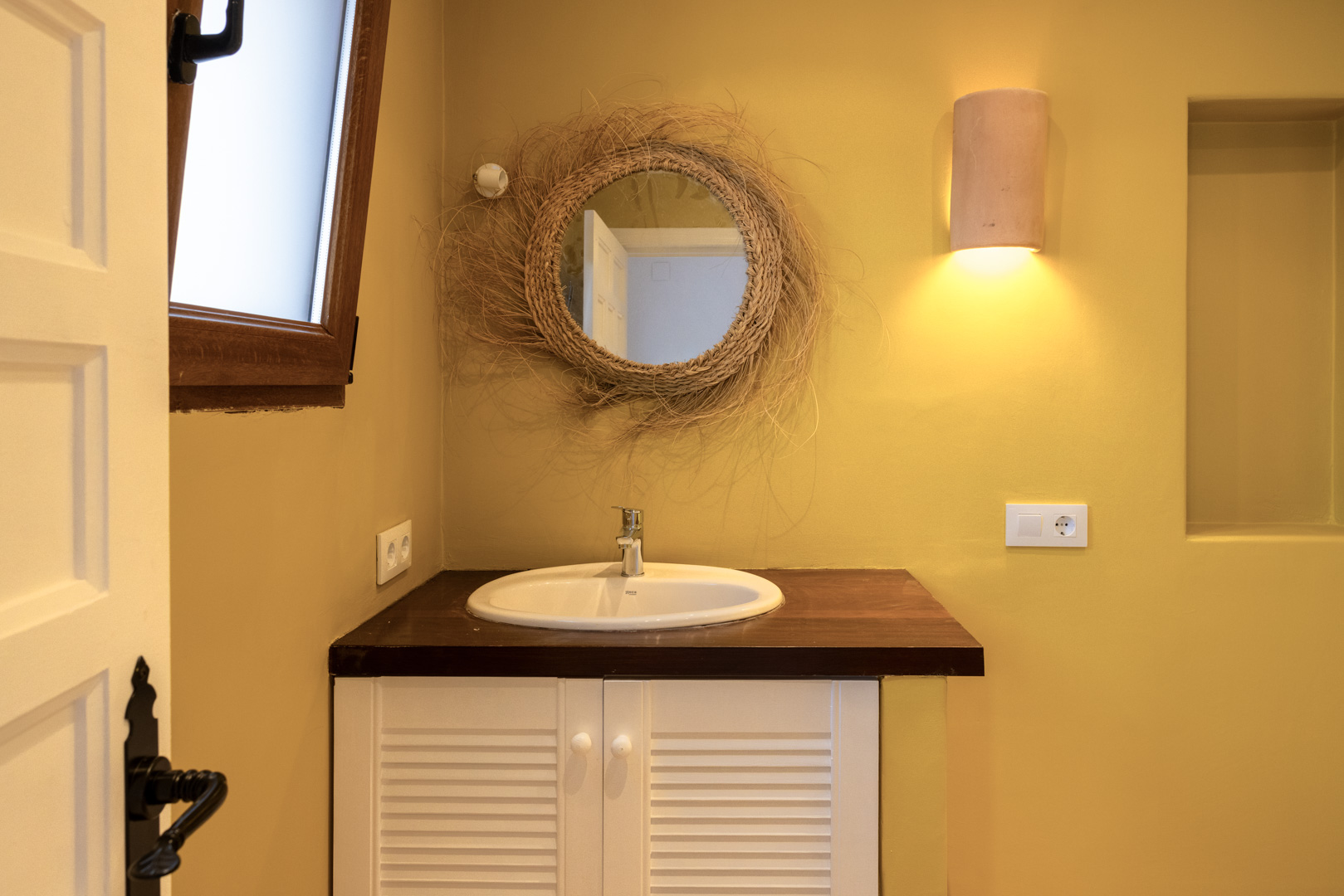 Detalle de baño con lavabo ovalado, espejo de mimbre y encimera de madera oscura sobre mueble blanco.