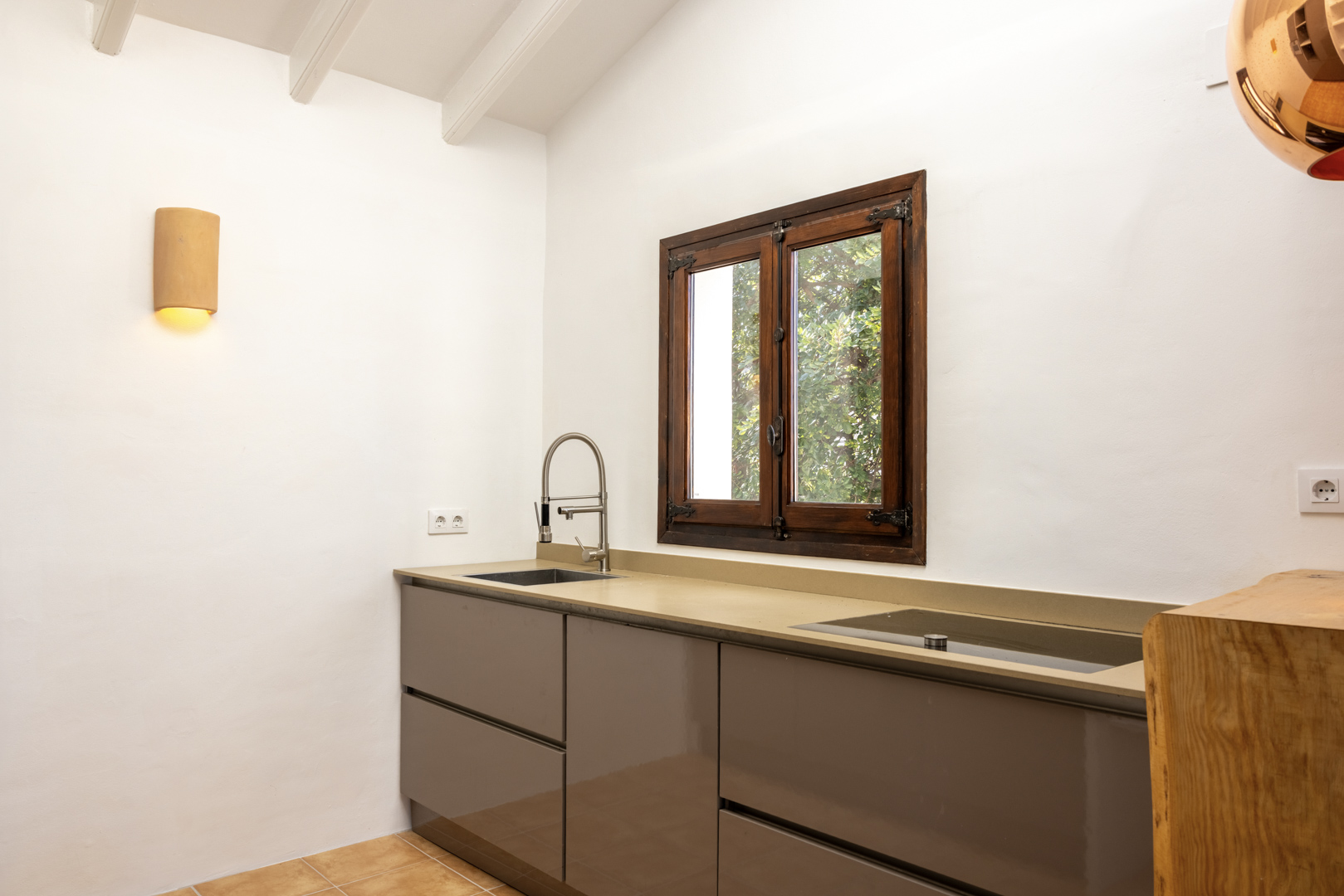 Detalle de cocina con encimera beige, fregadero de acero y ventana de madera con vistas al exterior.