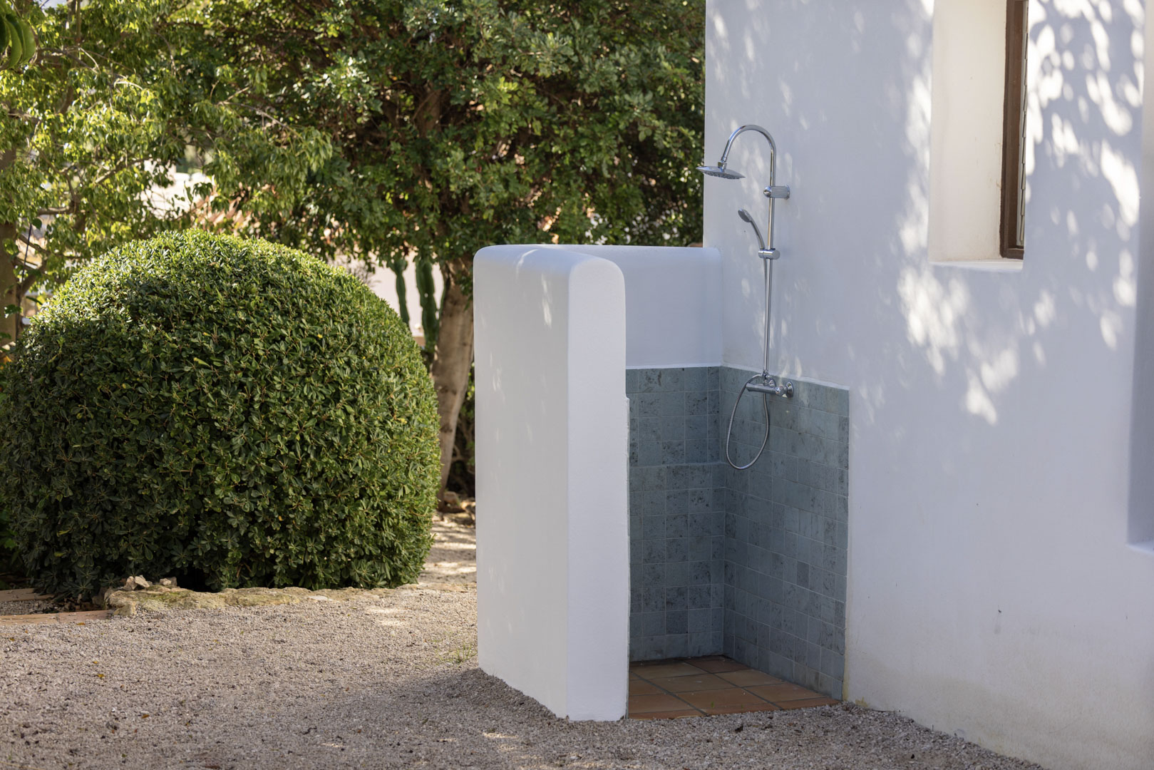 Detalle de ducha exterior con azulejos grises y base de terracota, junto a jardín con arbusto esférico.