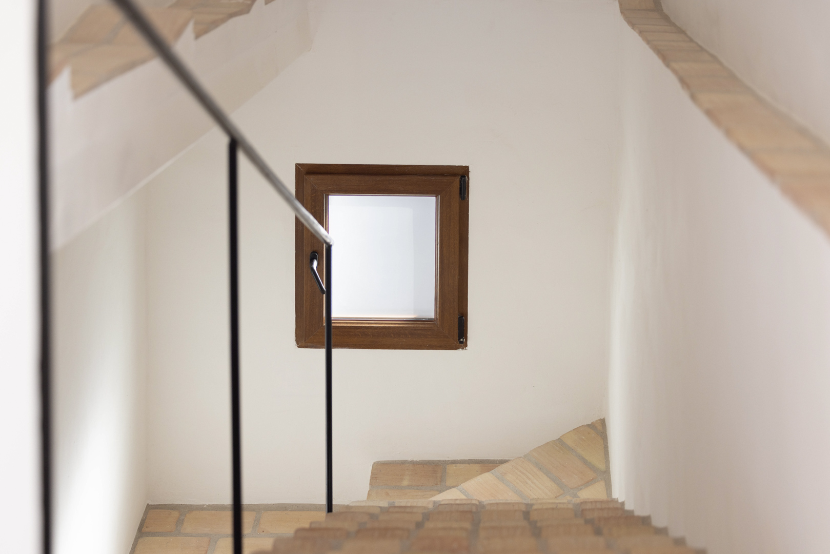 Detalle de escalera con barandilla negra y ventana de madera en pared blanca.