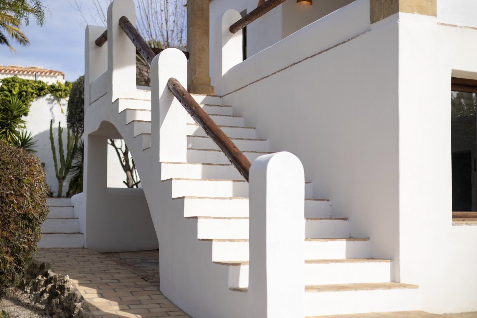 Detalle de escalera exterior blanca con barandilla de madera y pavimento de ladrillo.