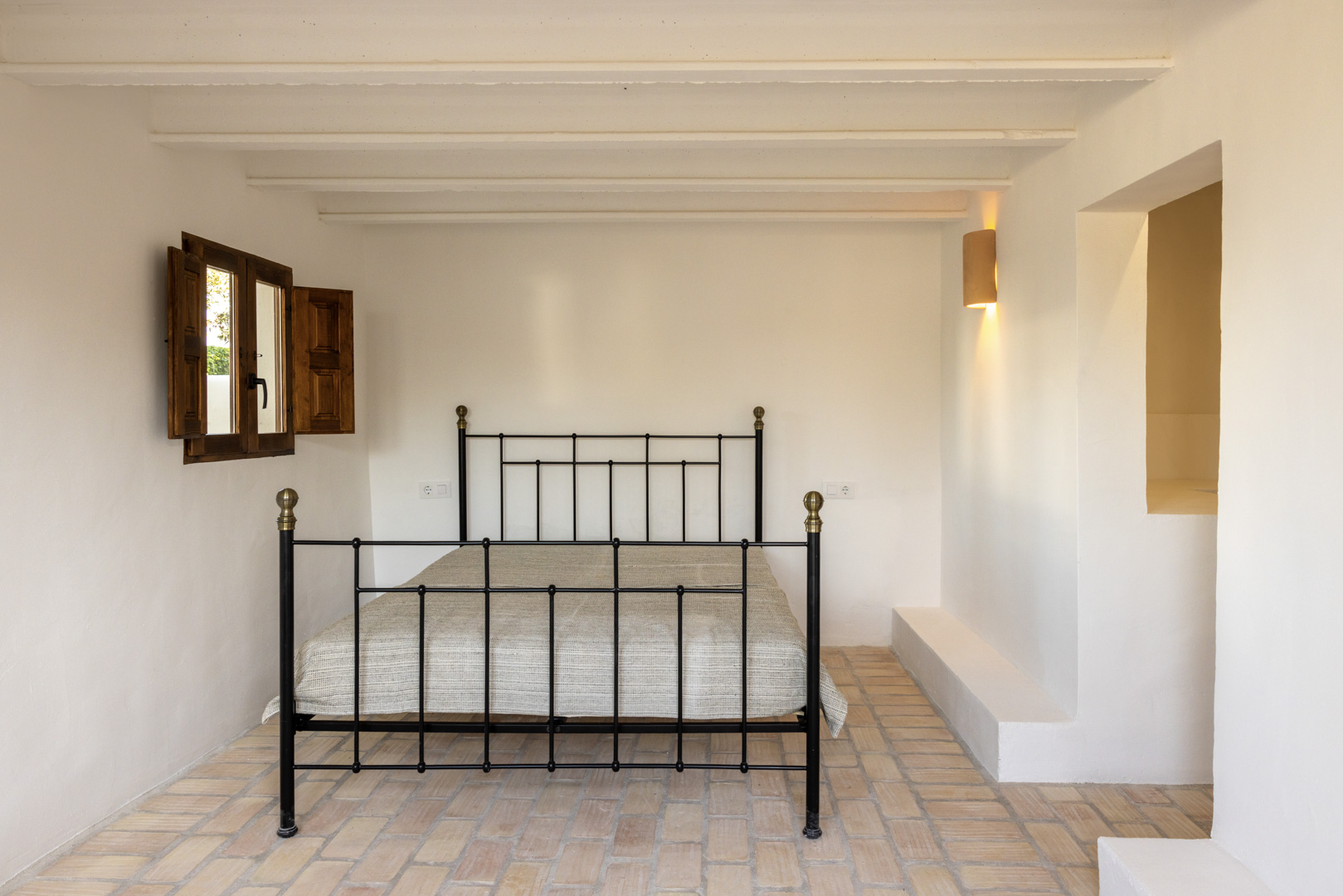 Dormitorio luminoso con cama de hierro forjado, contraventanas de madera y suelo de baldosas rústicas.