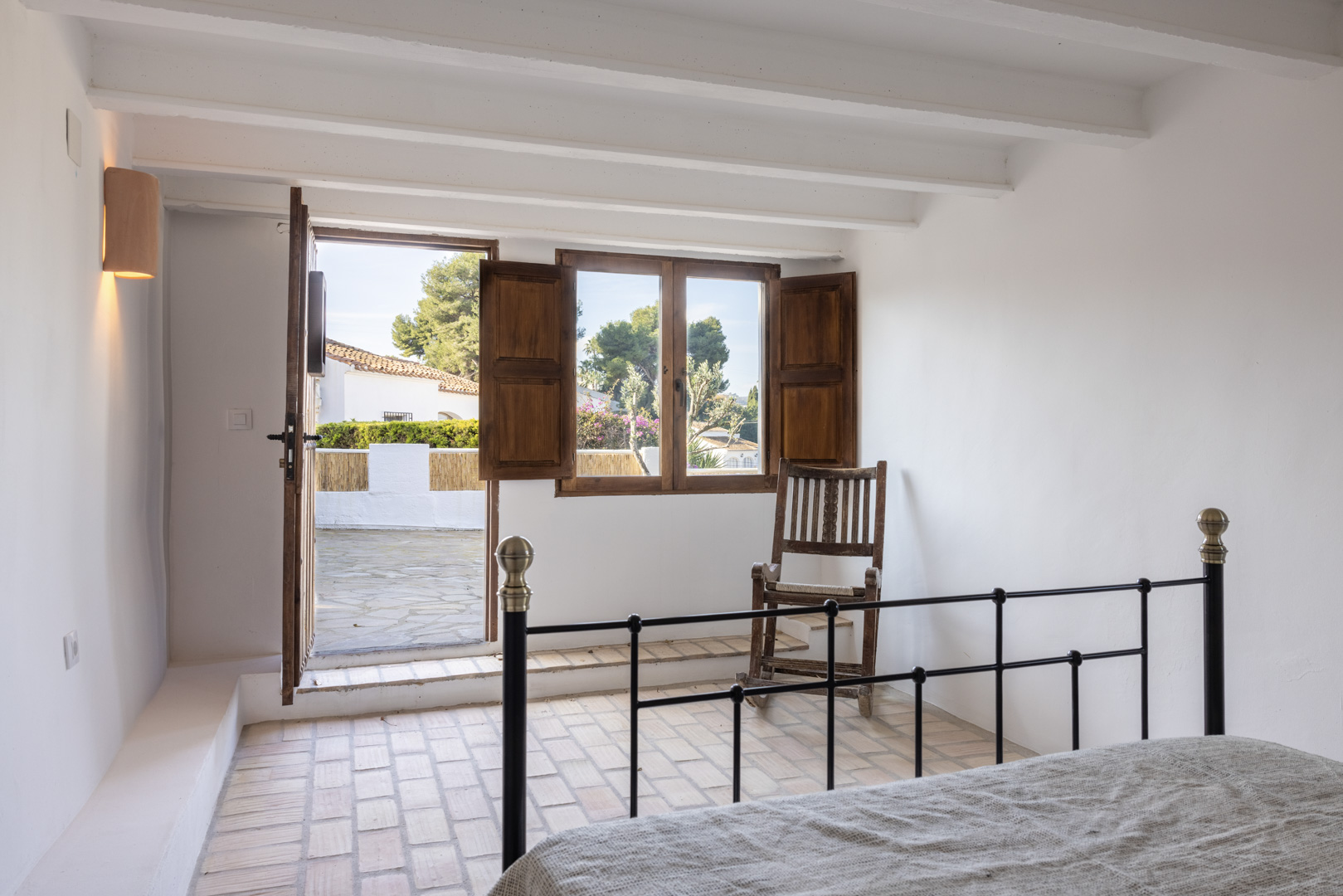 Dormitorio luminoso con suelo de terracota, puerta al patio y ventana con contraventanas de madera.