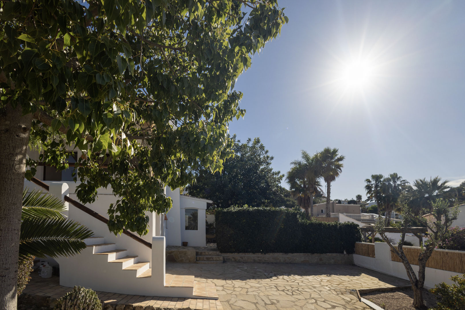 Exterior de la villa con patio de piedra, escaleras blancas y vegetación exuberante bajo el sol.