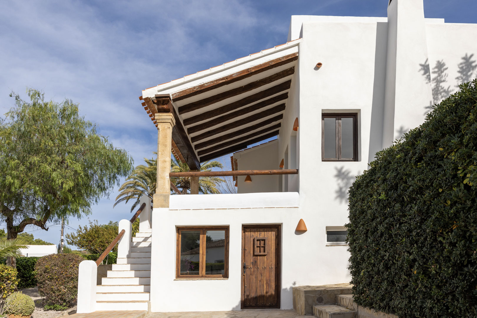 Fachada de villa mediterránea con porche de vigas de madera y acceso escalonado al jardín.