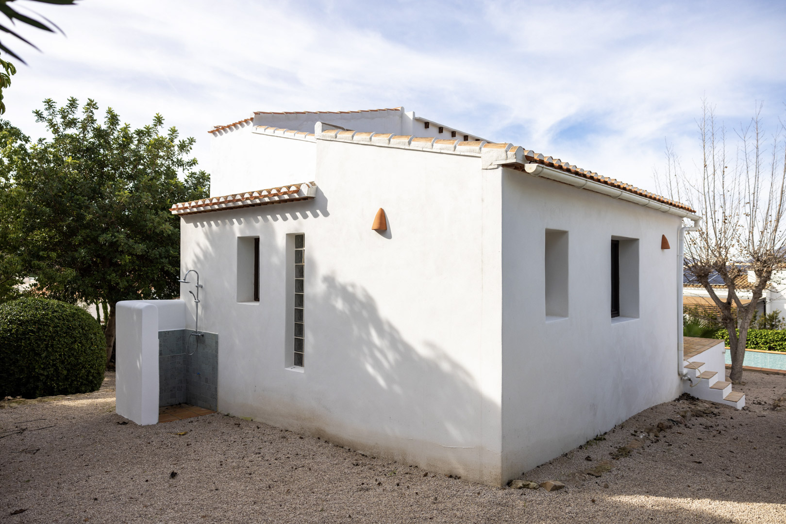 Vista exterior de la villa mostrando la ducha exterior con azulejos y la fachada blanca minimalista con ventanas.