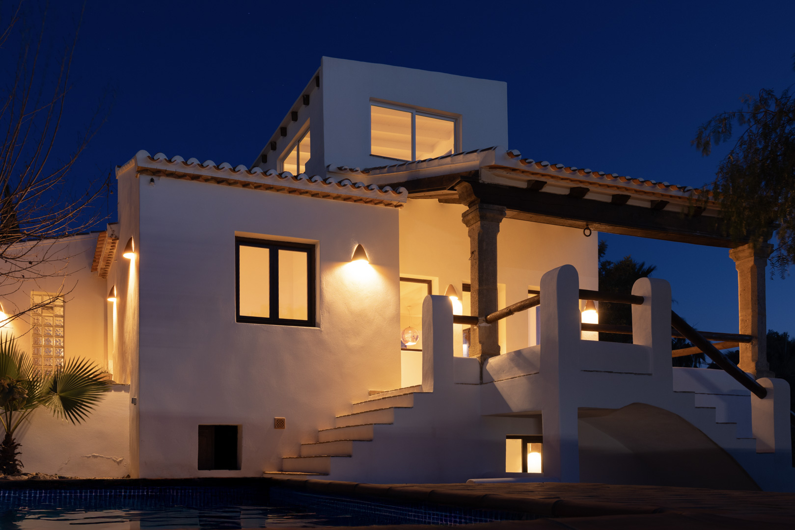Vista nocturna de la villa iluminada, destacando la escalera exterior y la terraza cubierta con columnas de piedra.