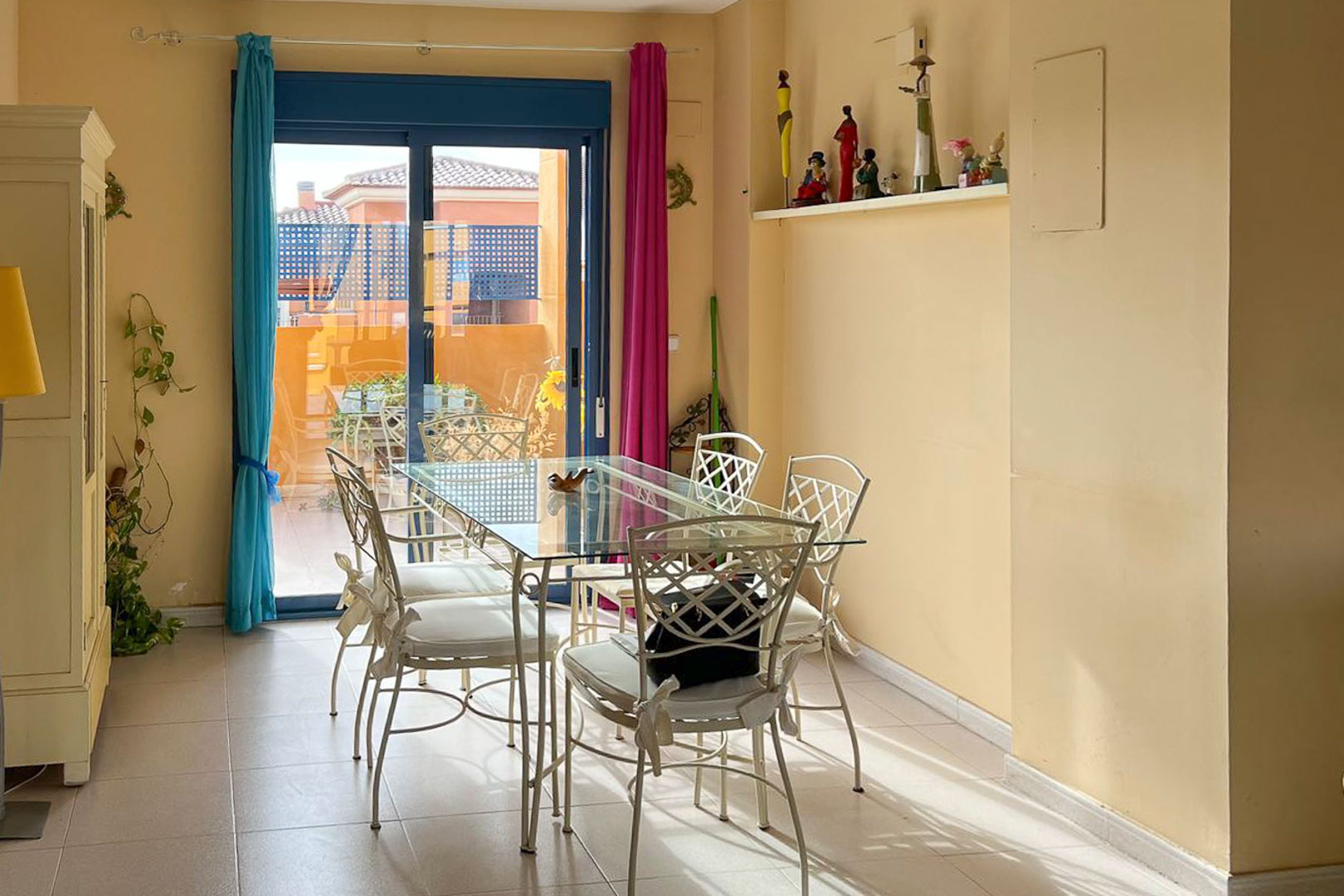 Comedor luminoso con mesa de cristal y sillas de hierro forjado, acceso a terraza con vistas.