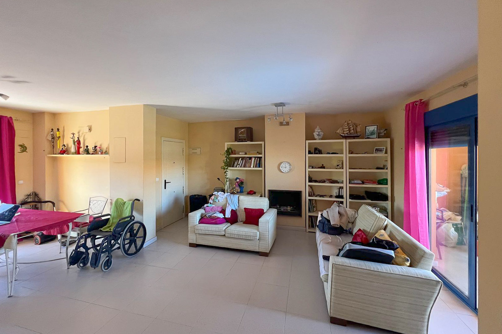 Salón comedor con estanterías, sofás tapizados y acceso a terraza con cortinas rosas.