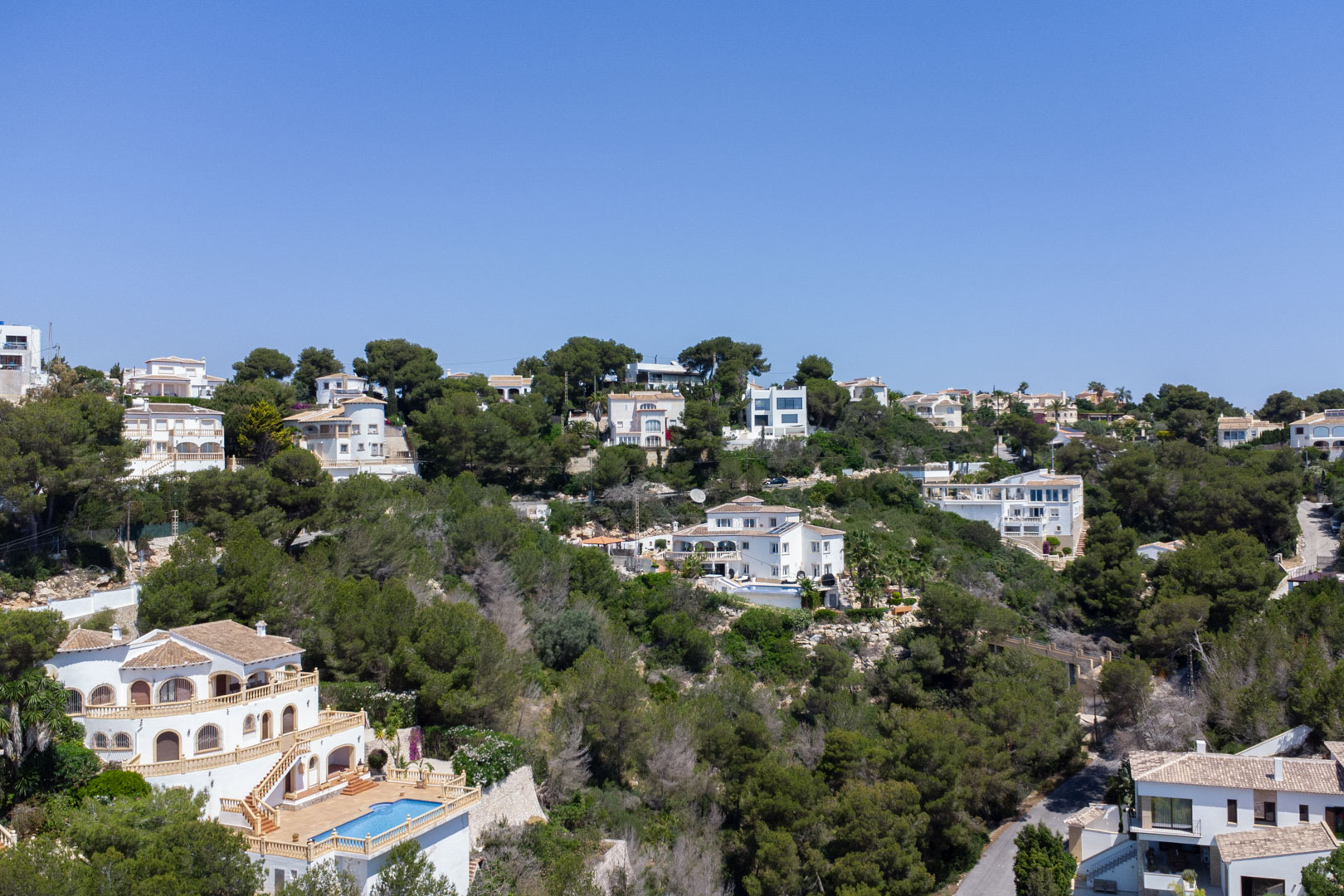 Vista panorámica de la urbanización Costa Nova en Jávea, con villas mediterráneas integradas en una ladera boscosa de pinos bajo un cielo despejado.