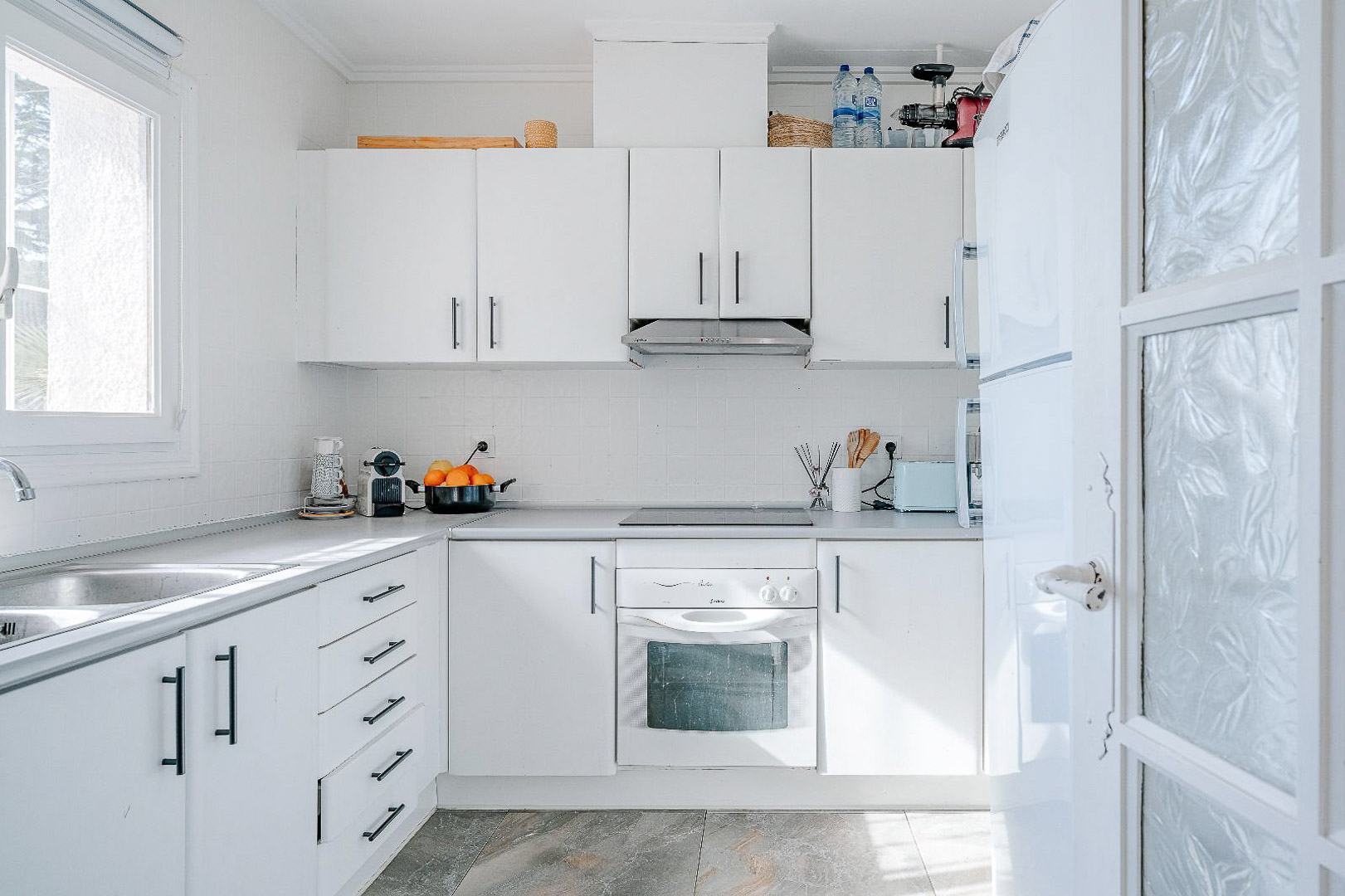 Cocina funcional en tonos blancos con tiradores negros, equipada con horno integrado, placa vitrocerámica y fregadero de doble seno bajo una ventana con luz natural.