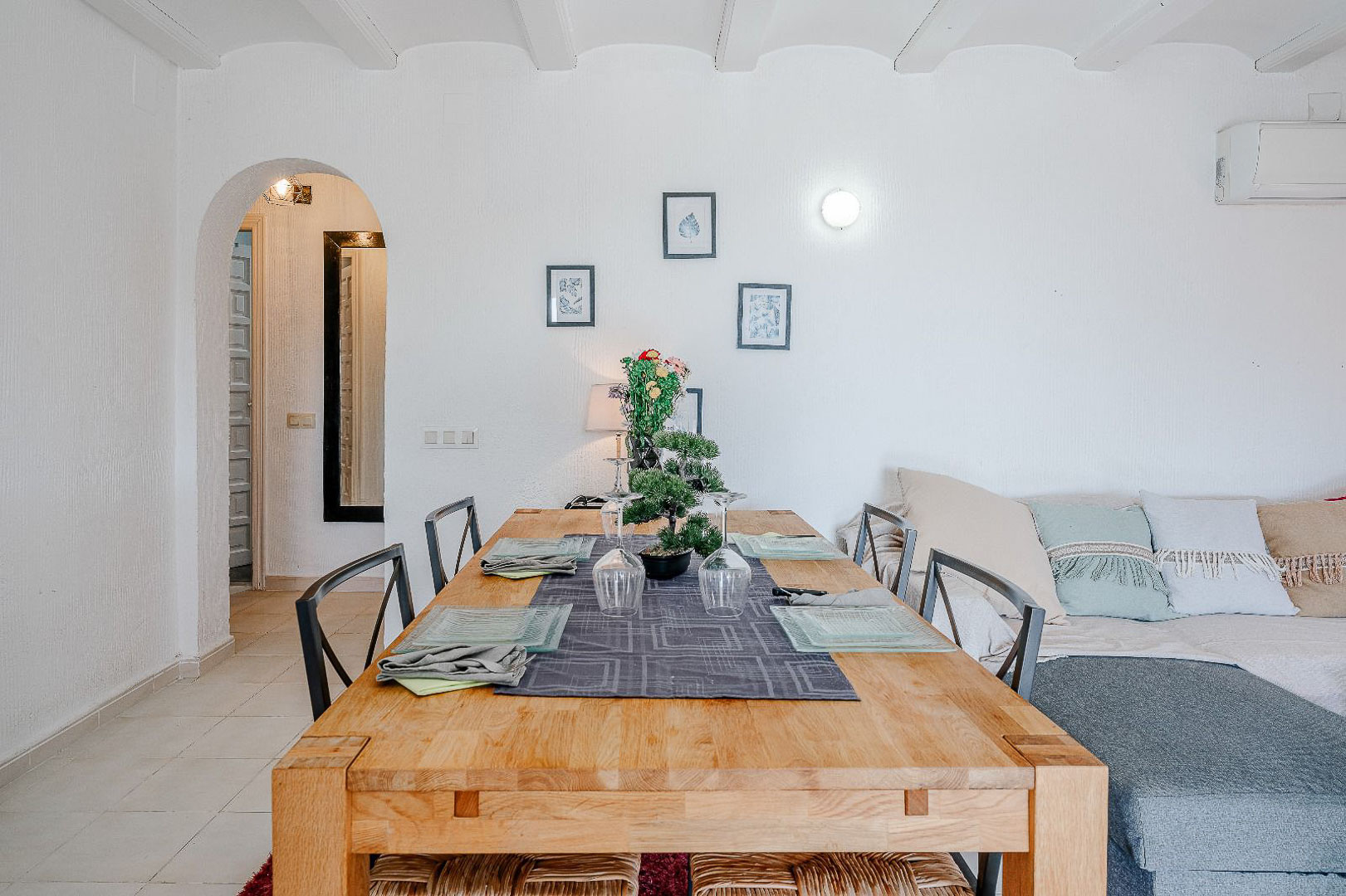 Comedor luminoso con mesa de madera maciza y techo de vigas blancas. Un arco conecta con el pasillo junto a la zona de estar con sofá y aire acondicionado.
