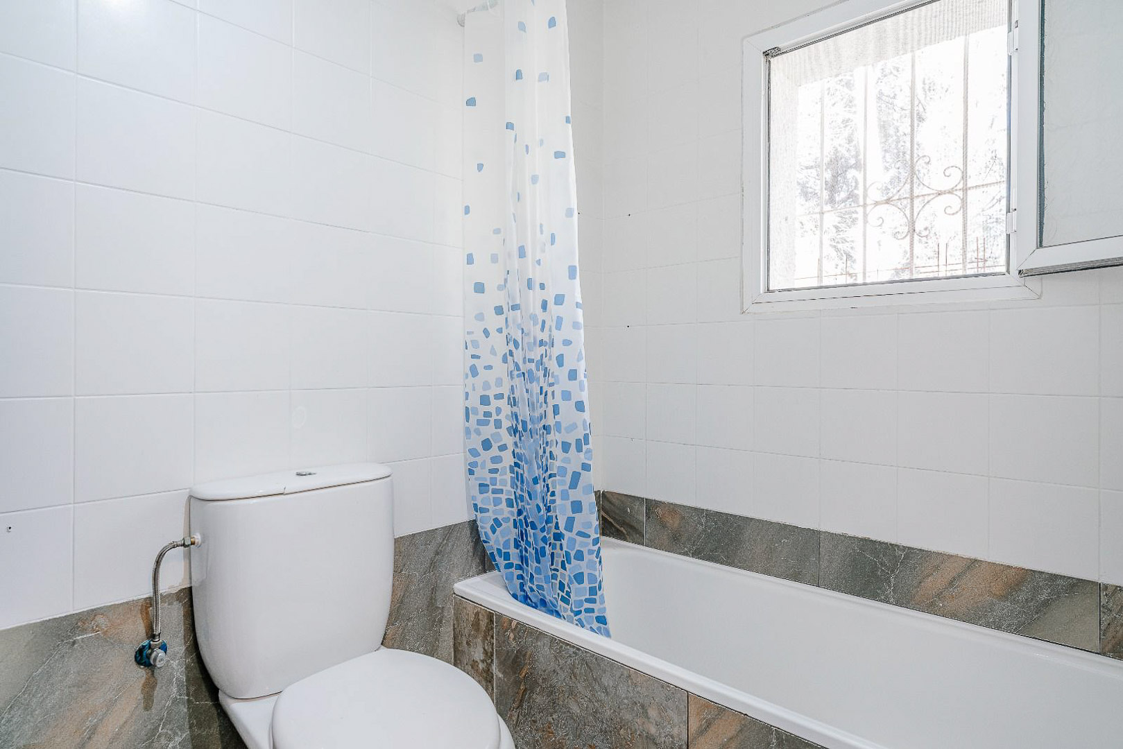 Cuarto de baño renovado con bañera, inodoro de cerámica blanca y azulejos en tonos neutros. Una ventana exterior con reja aporta luz natural y ventilación.