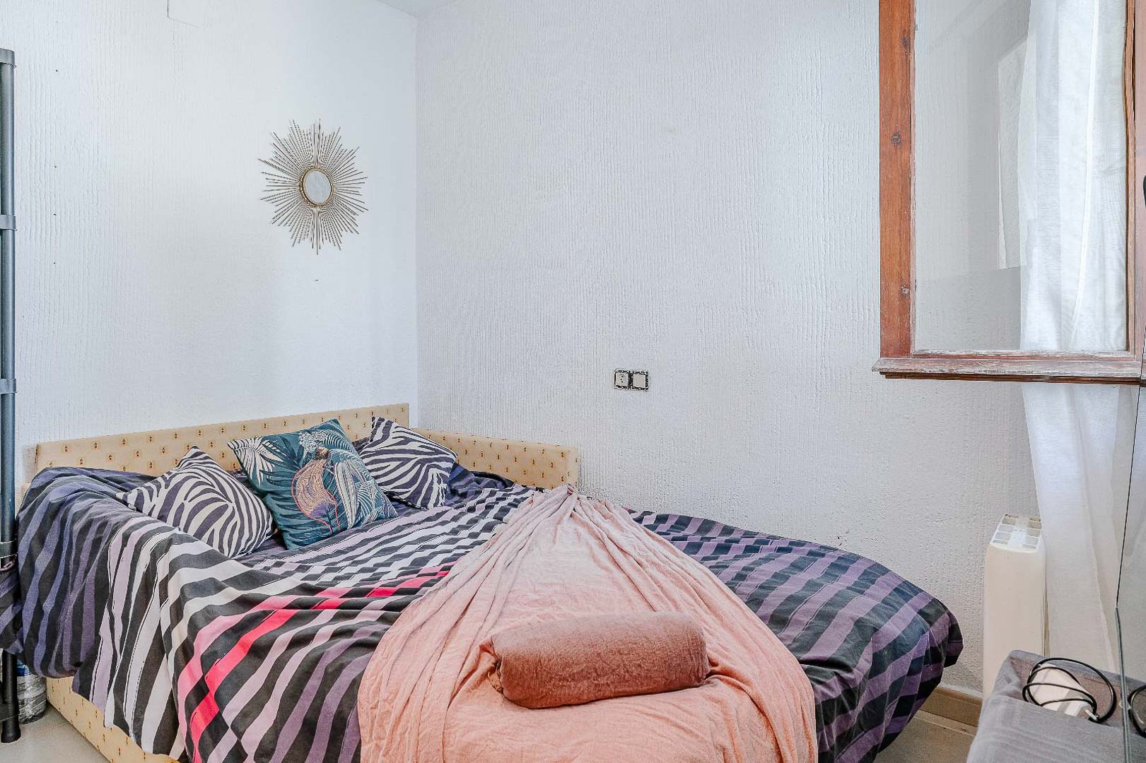 Dormitorio luminoso con paredes de acabado rugoso, cama individual con cabecero tapizado y ventana de madera que aporta luz natural y ventilación.