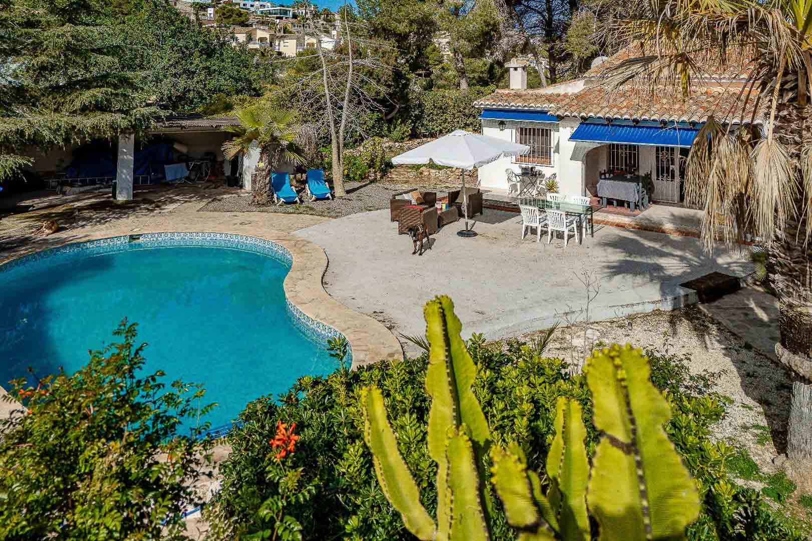 Exterior con piscina privada de forma irregular, amplia terraza pavimentada y fachada blanca con toldos azules rodeada de vegetación mediterránea.