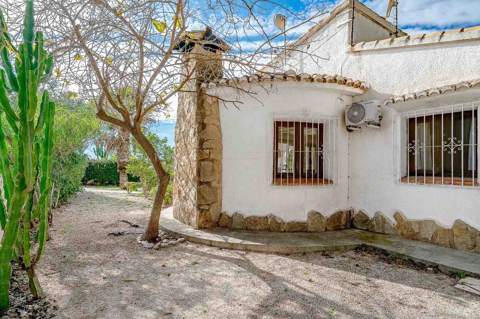 Exterior de estilo mediterráneo con fachada blanca, chimenea de piedra natural, ventanas de madera con rejas decorativas y un jardín de grava con vegetación autóctona.