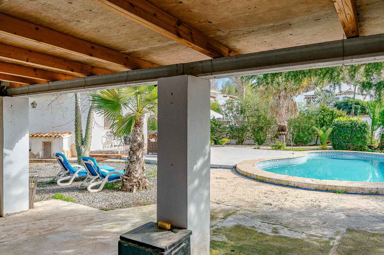 Porche cubierto con vigas de madera vista que ofrece vistas directas a la piscina de forma riñonada y al jardín mediterráneo con palmeras.