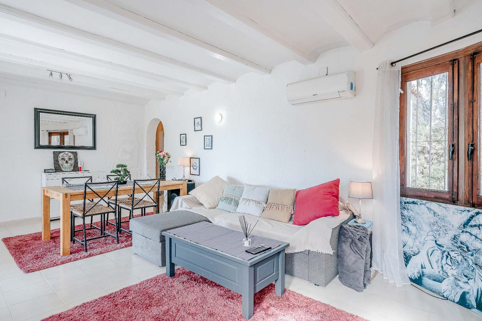 Salón comedor de concepto abierto con techos de vigas blancas, aire acondicionado tipo split, suelos cerámicos y una zona de comedor integrada con mobiliario de madera.