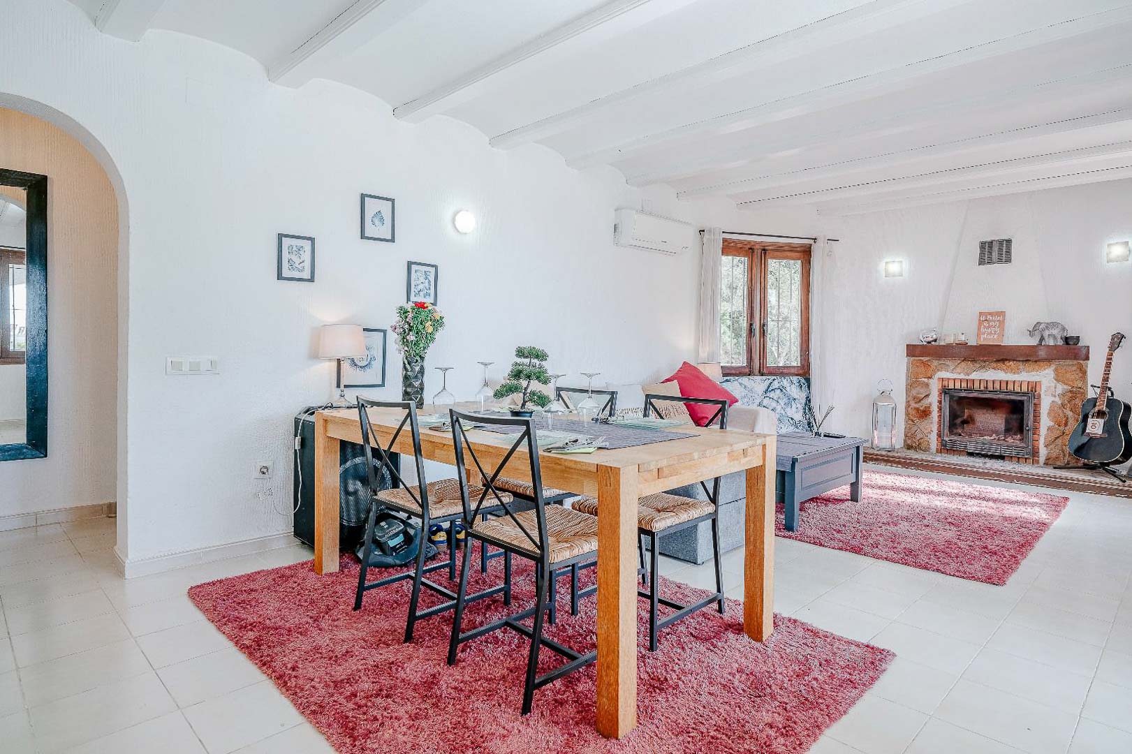 Salón comedor de concepto abierto con techos de vigas blancas, chimenea de piedra natural y zona de comedor con mesa de madera sobre alfombras decorativas.
