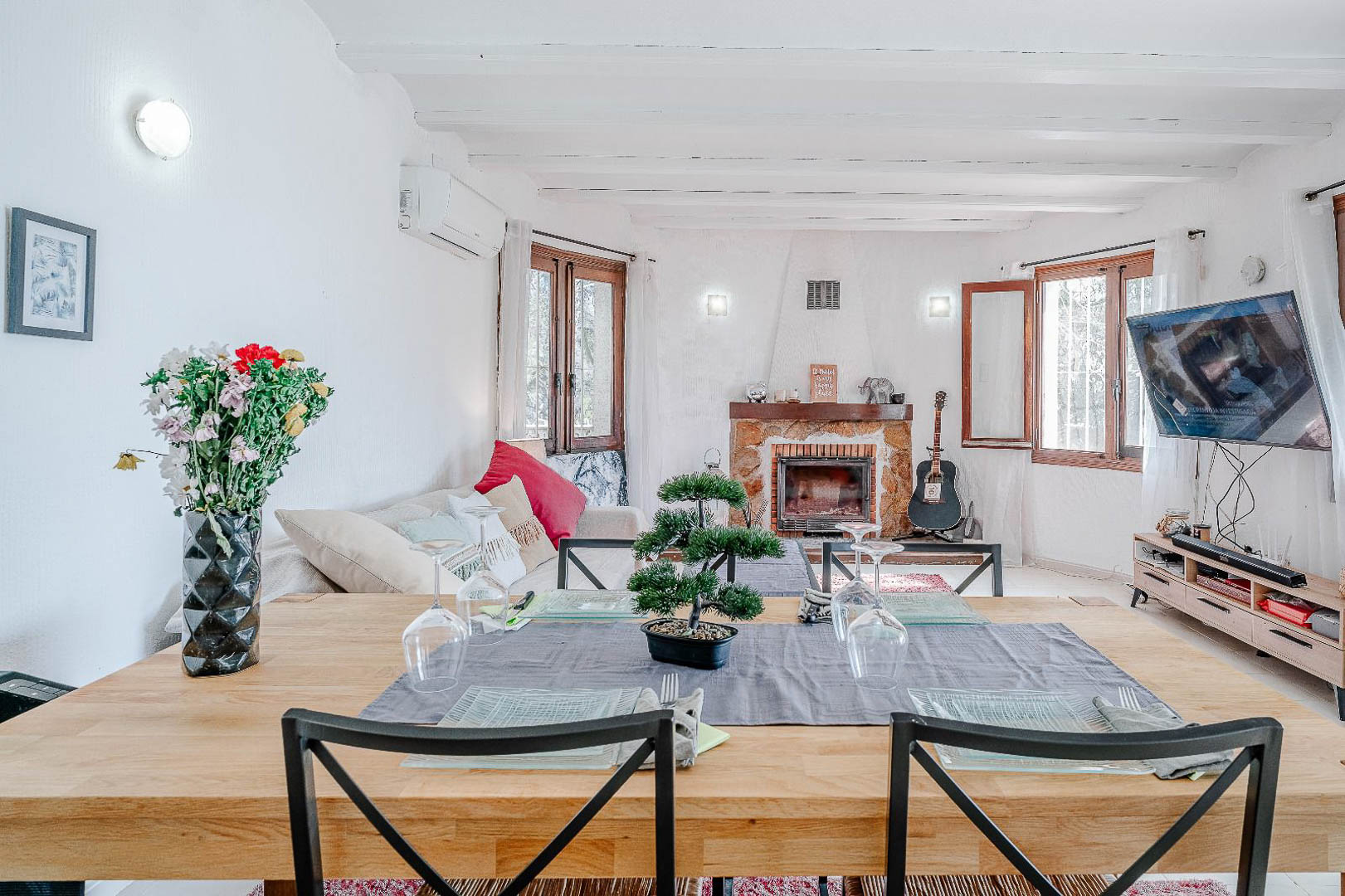 Salón comedor de planta abierta con vigas vistas en blanco, chimenea de piedra rústica y ventanales de madera que aportan gran luminosidad al espacio.